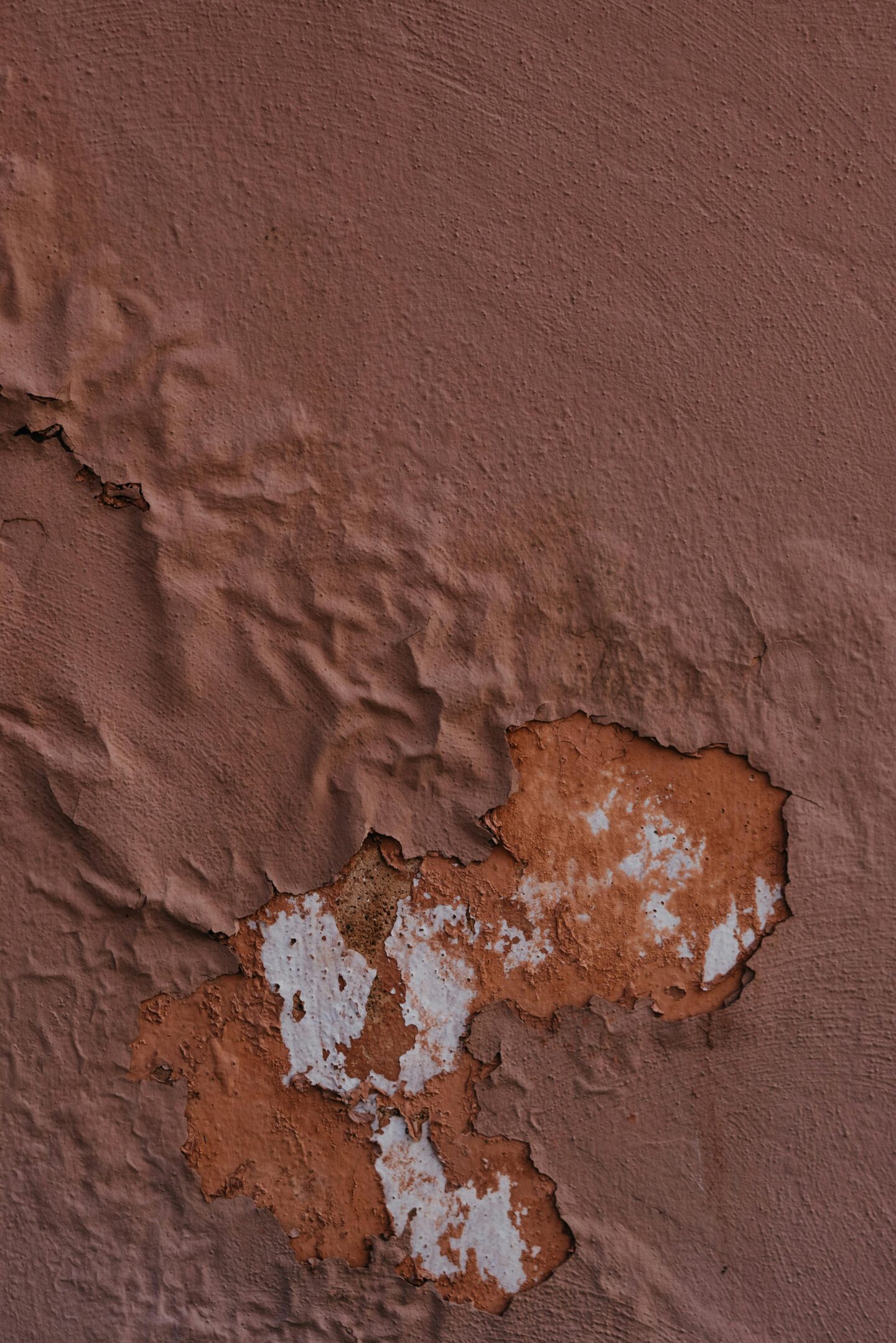 Close-up of a wall with reddish-brown paint that is peeling and bubbling, revealing layers of lighter paint and bare surface underneath.