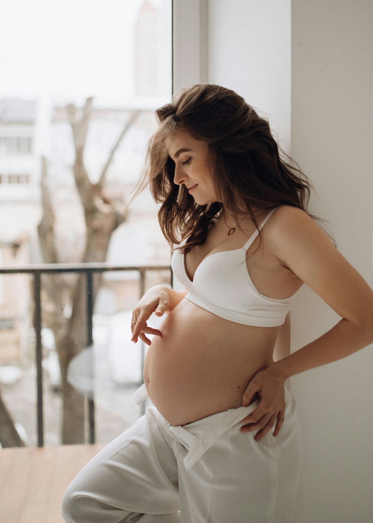 Smiling pregnant woman in a white bra and loose white pants gently touching her belly while standing by a bright window.
