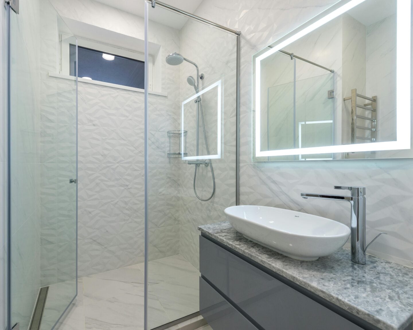 Modern bathroom with a gray quartz countertop, white vessel sink, lighted mirror, and glass-enclosed shower with textured white tile.