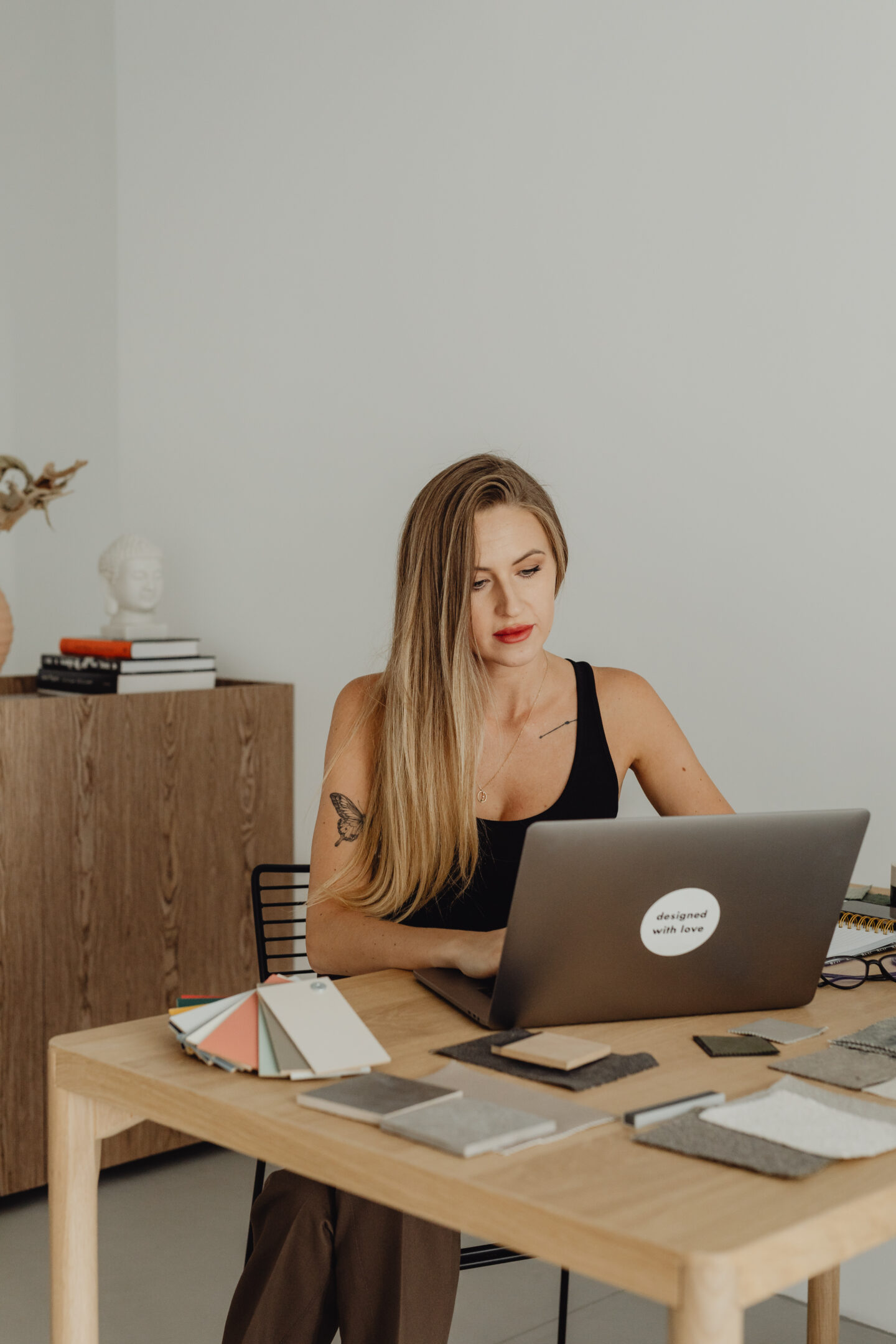 Woman working on a laptop at a wooden desk with fabric samples and color swatches.