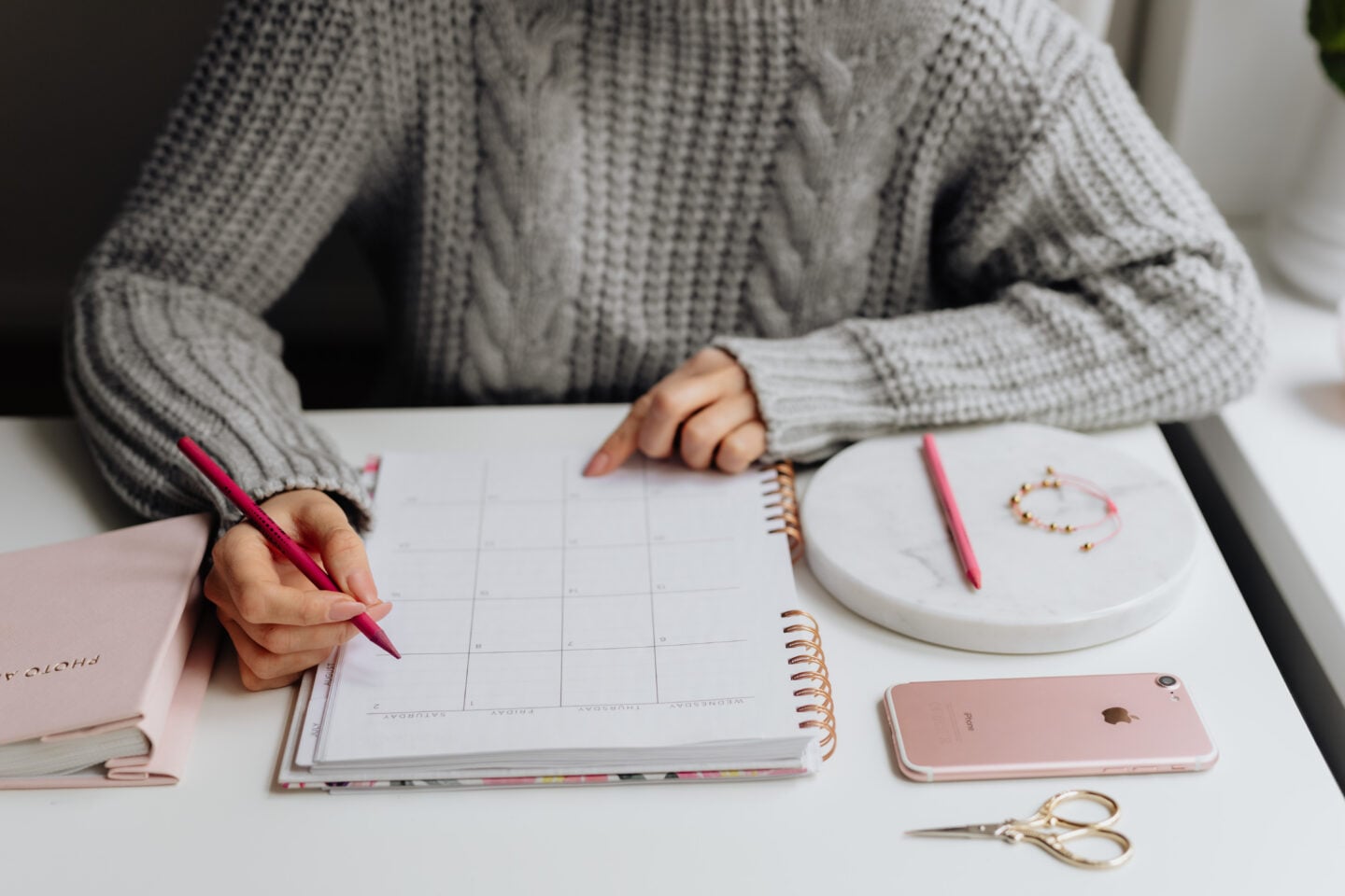 Woman in a cozy grey sweater writing in a calendar planner with a pink pen, symbolizing organizing daily schedules and keeping track of today’s date.