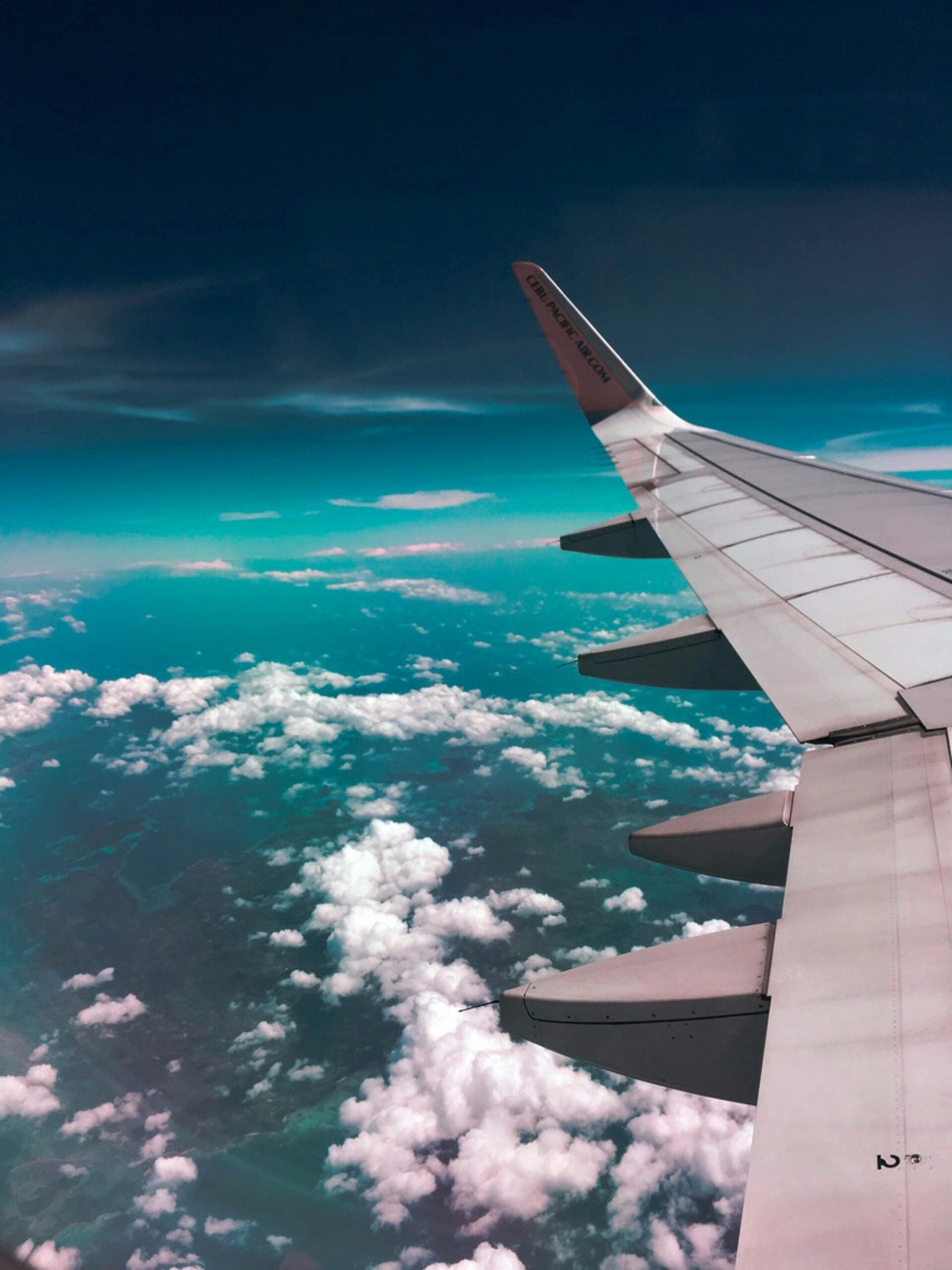 View from an airplane window showing the aircraft wing above clouds and turquoise sky during flight.