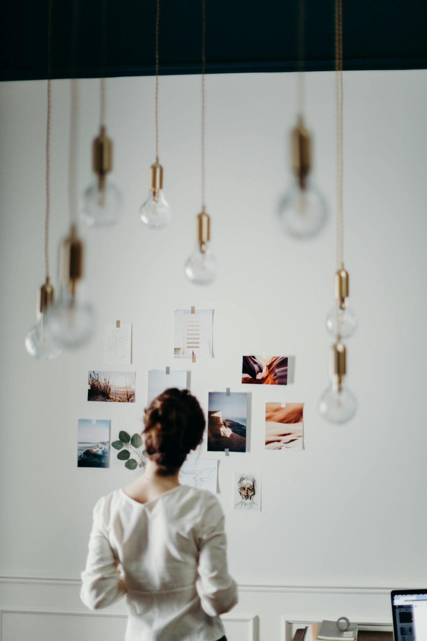 Woman standing in front of a wall covered with inspiration photos and art prints, with hanging light bulbs in the foreground.