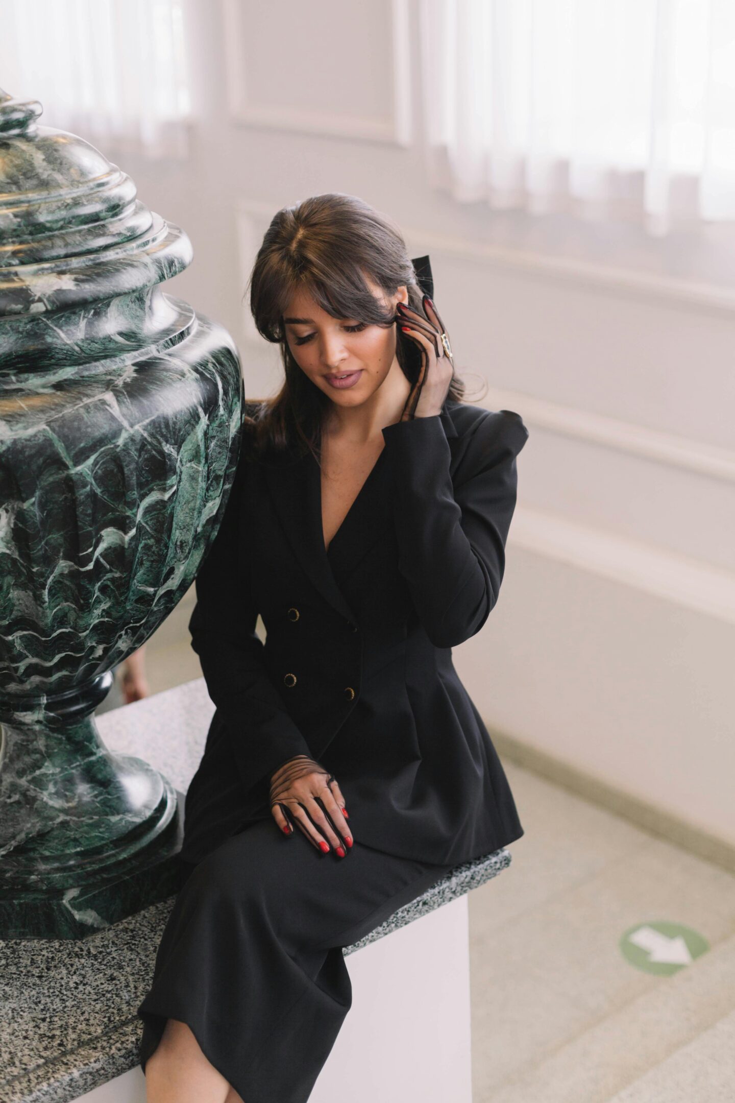 Elegant woman in a black blazer dress sitting indoors, touching her hair and smiling softly.