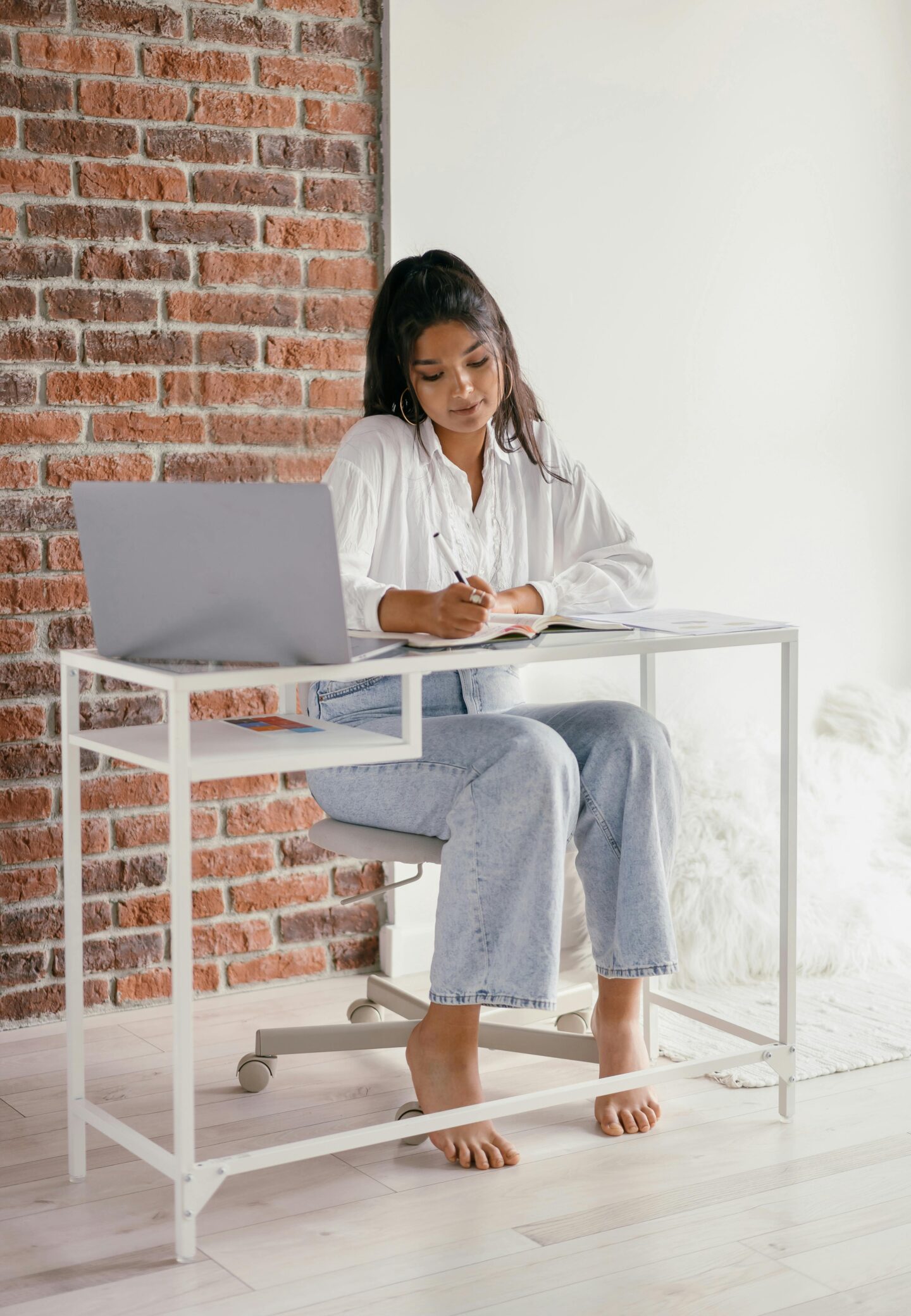 Young woman sitting barefoot at a desk with a laptop, writing in a notebook beside a brick wall.