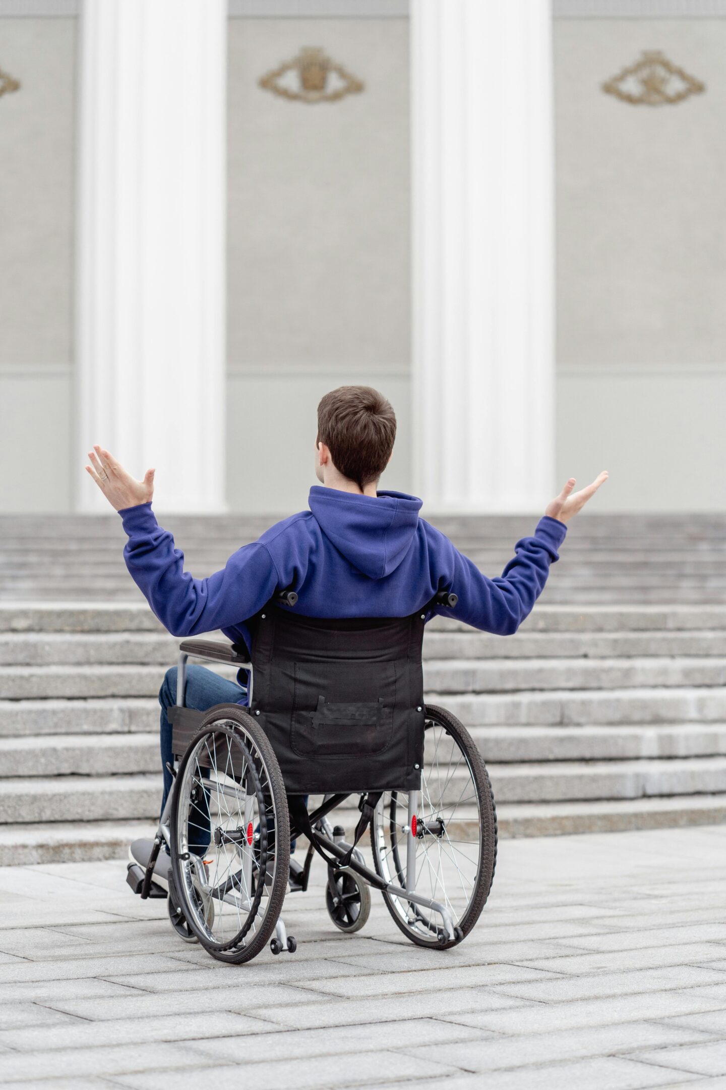 A person in a wheelchair faces a long flight of stairs with arms raised in frustration or disbelief, wearing a blue hoodie in front of a building with large white columns.
