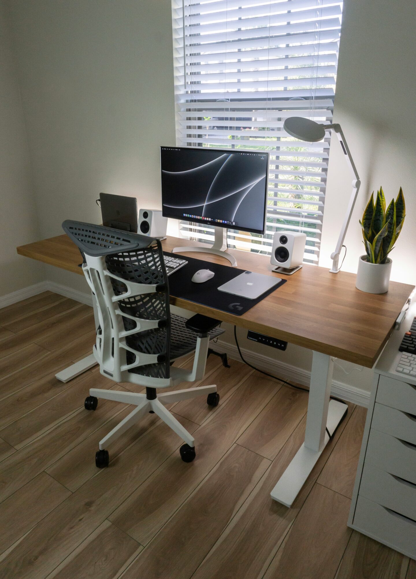 A modern home office setup featuring an ergonomic white office chair, a wooden desk with a computer monitor, laptop, speakers, desk lamp, and a potted plant near a window with blinds.