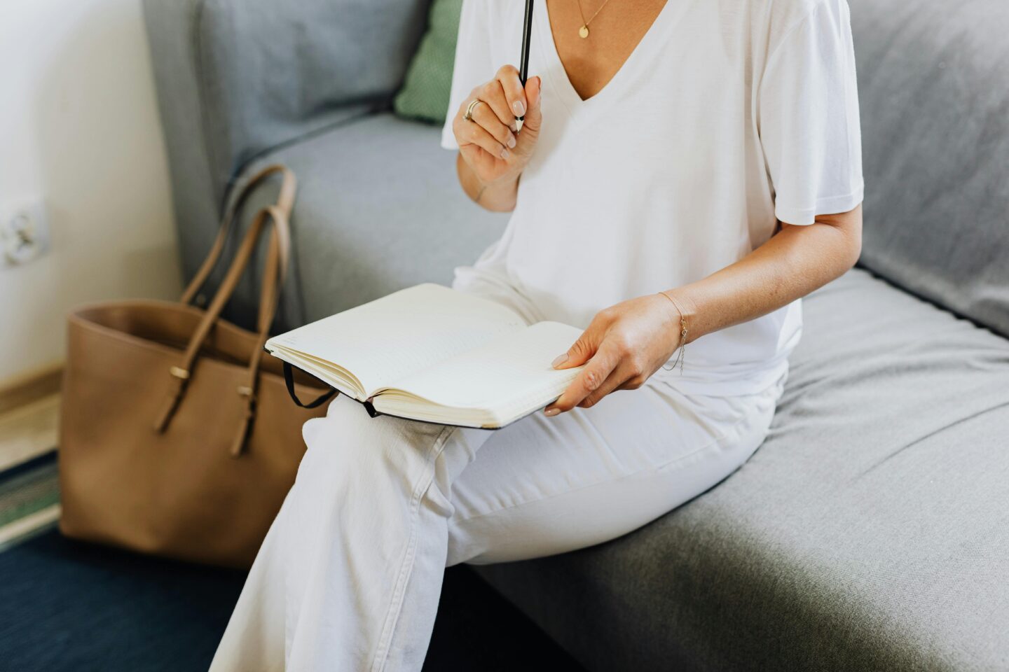 Person sitting on a couch in white clothes, holding an open notebook and pen, with a tan handbag on the floor nearby.