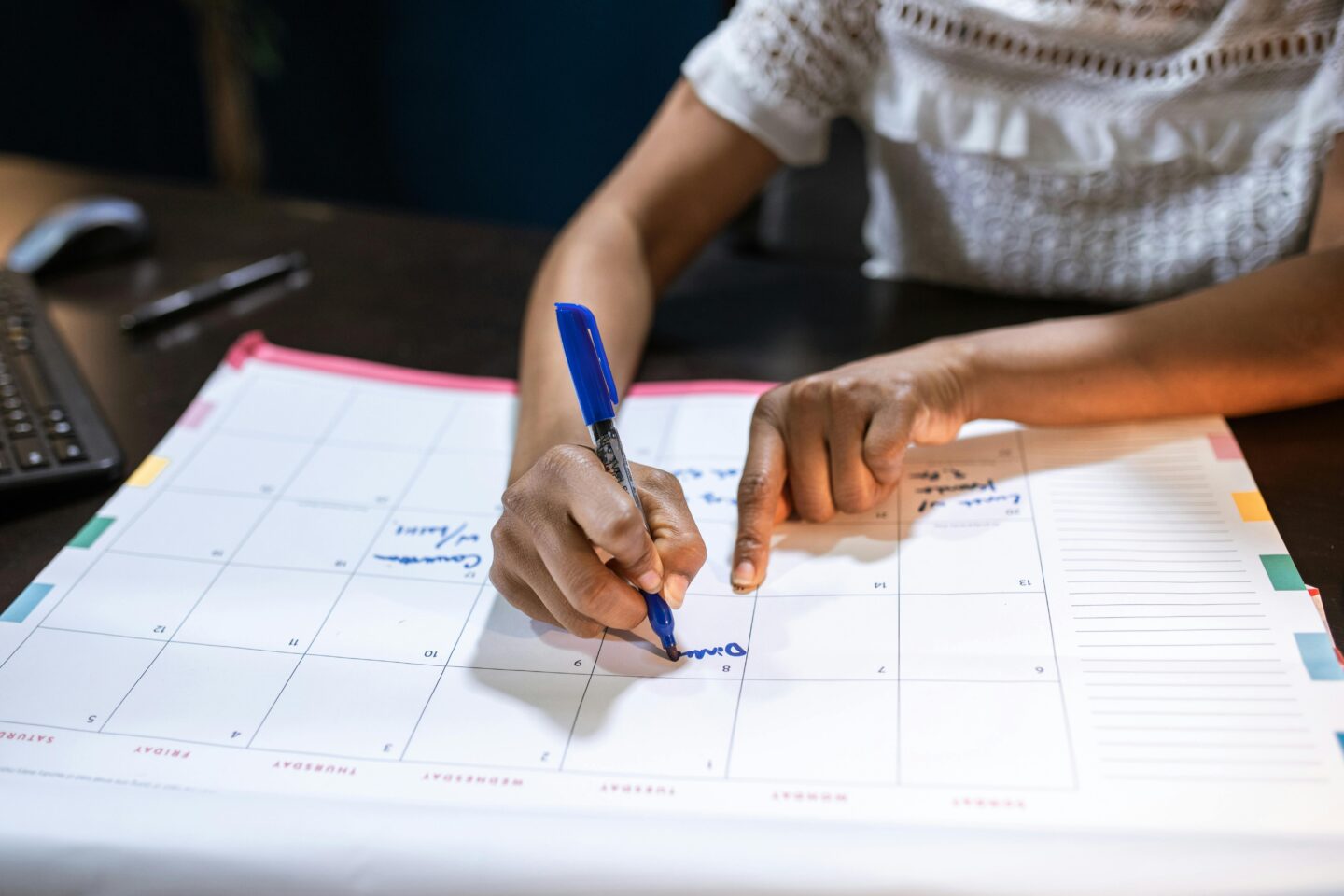 Person writing on a large desk calendar with a blue pen, planning their schedule.