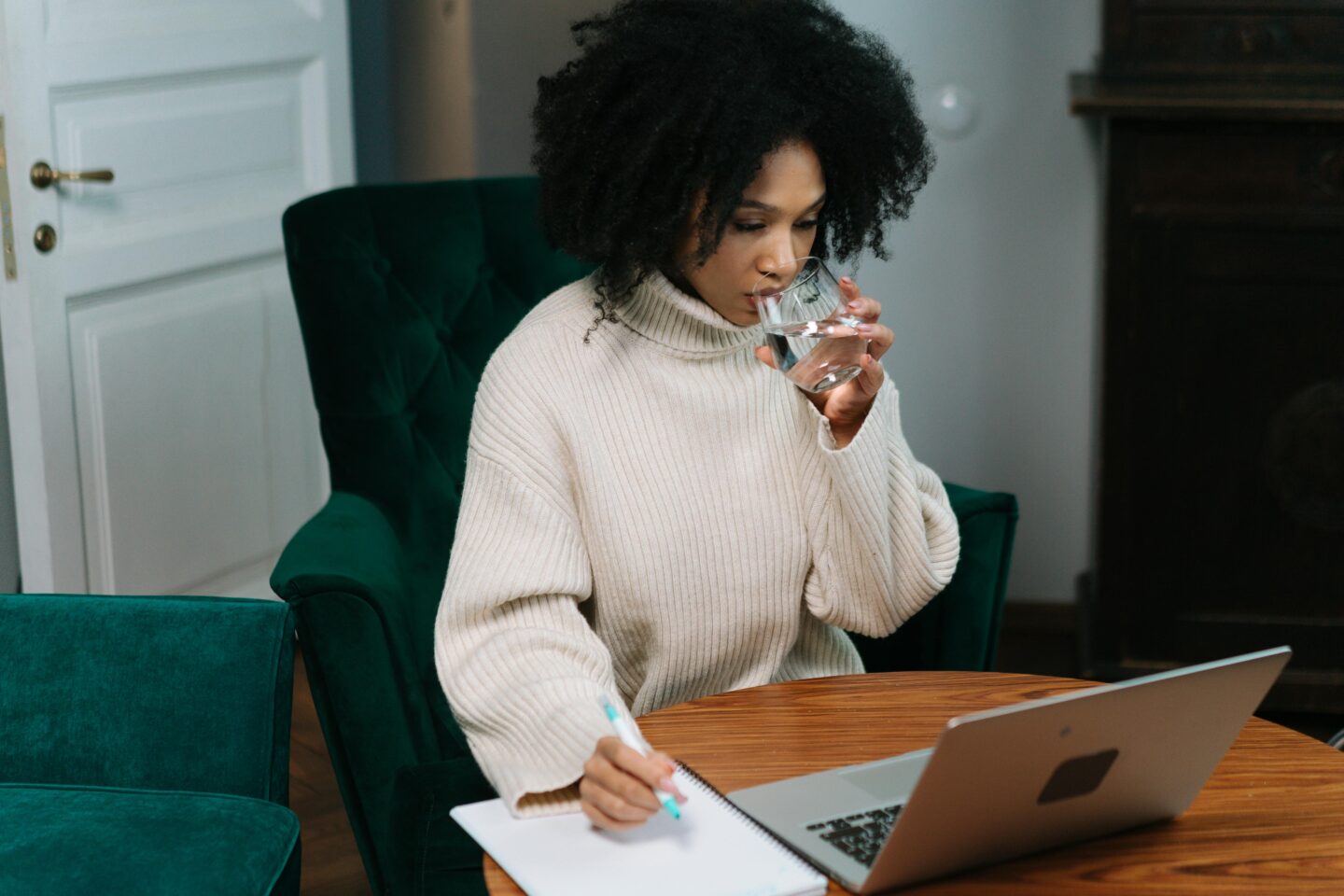 Woman sitting at a table with a laptop and notebook, organizing her schedule before Christmas while drinking a glass of water.