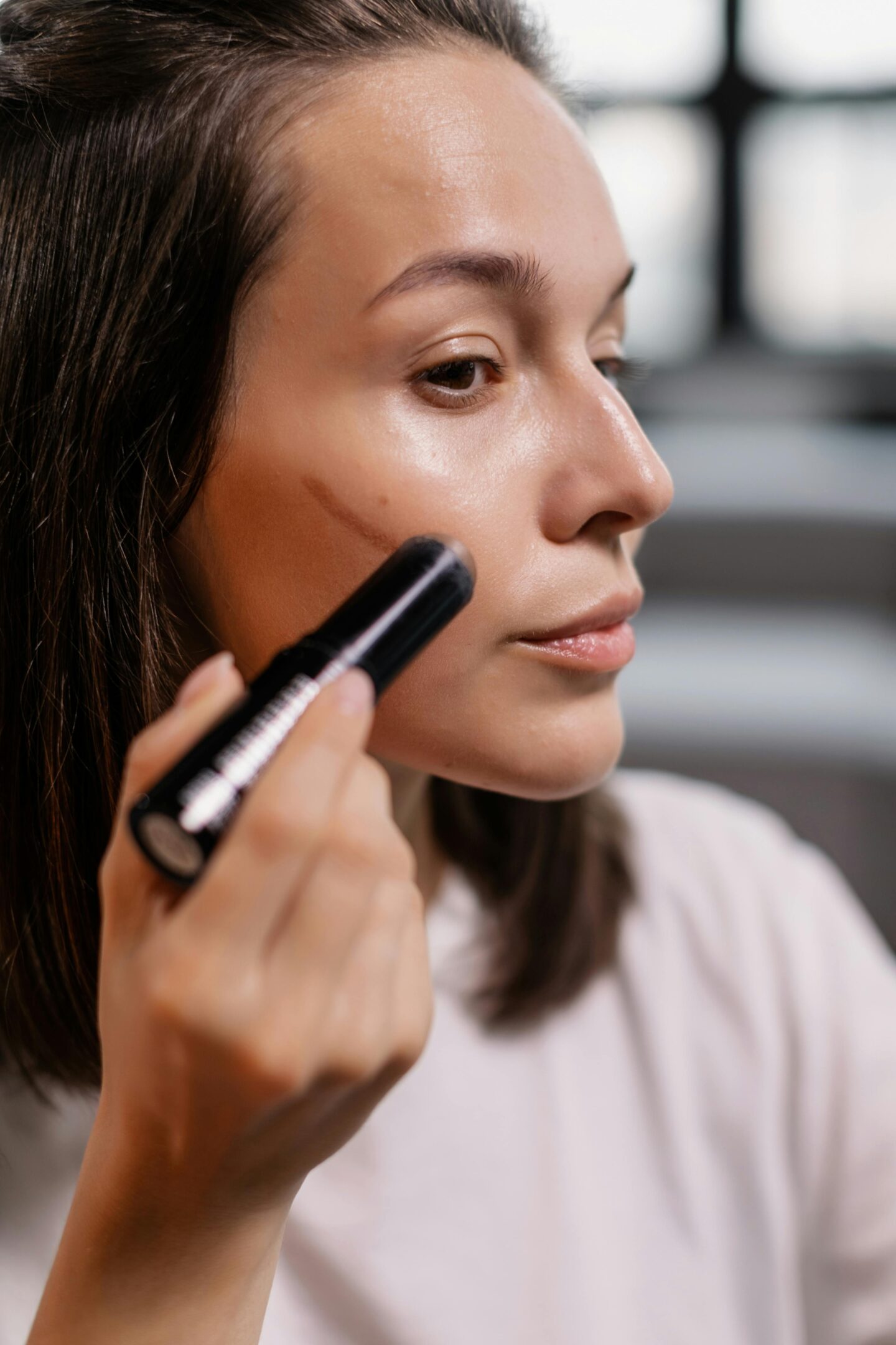 A woman applies contour makeup stick along her cheekbone.