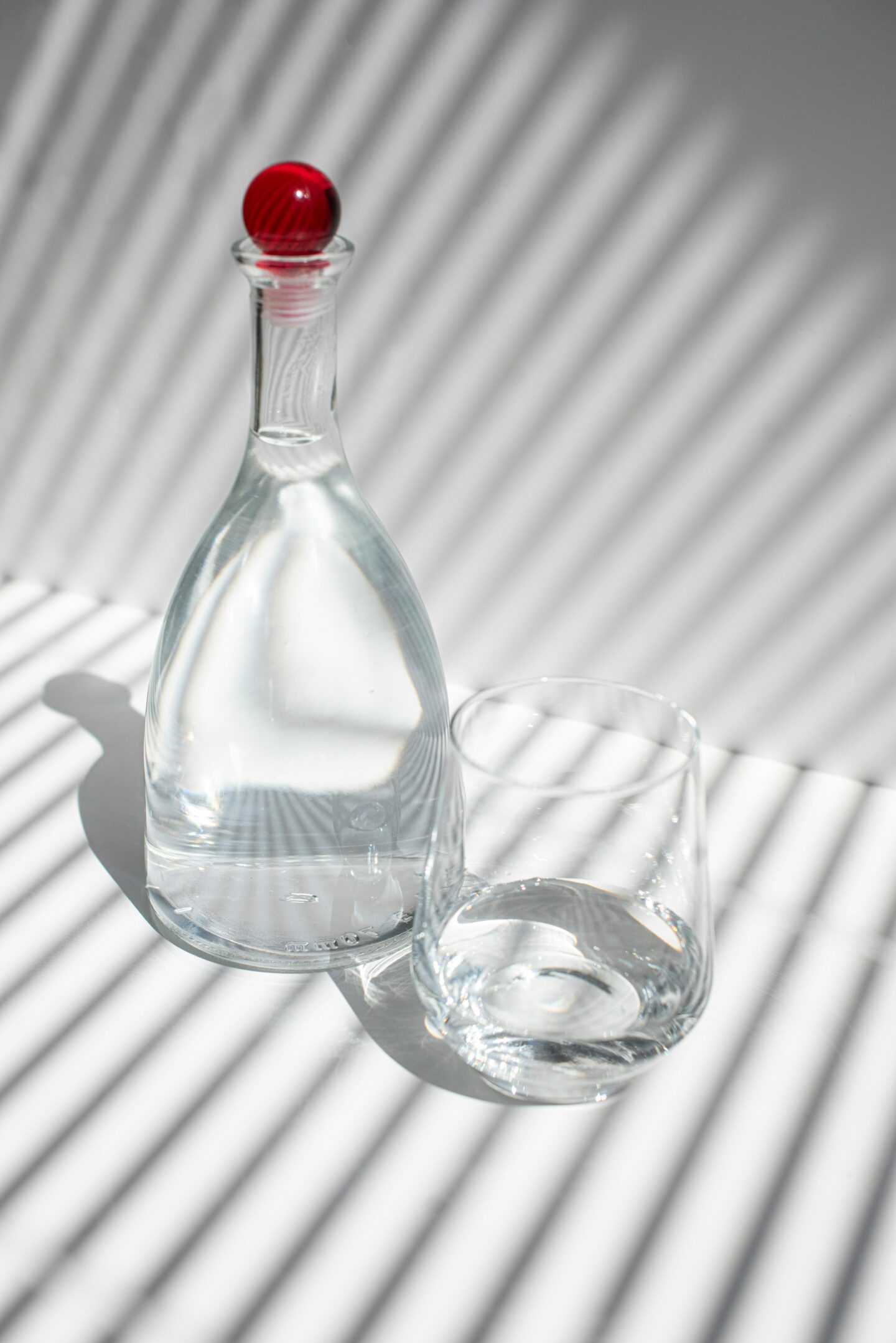 A clear glass bottle with a red cap and a glass of water placed on a white surface with striped shadows.