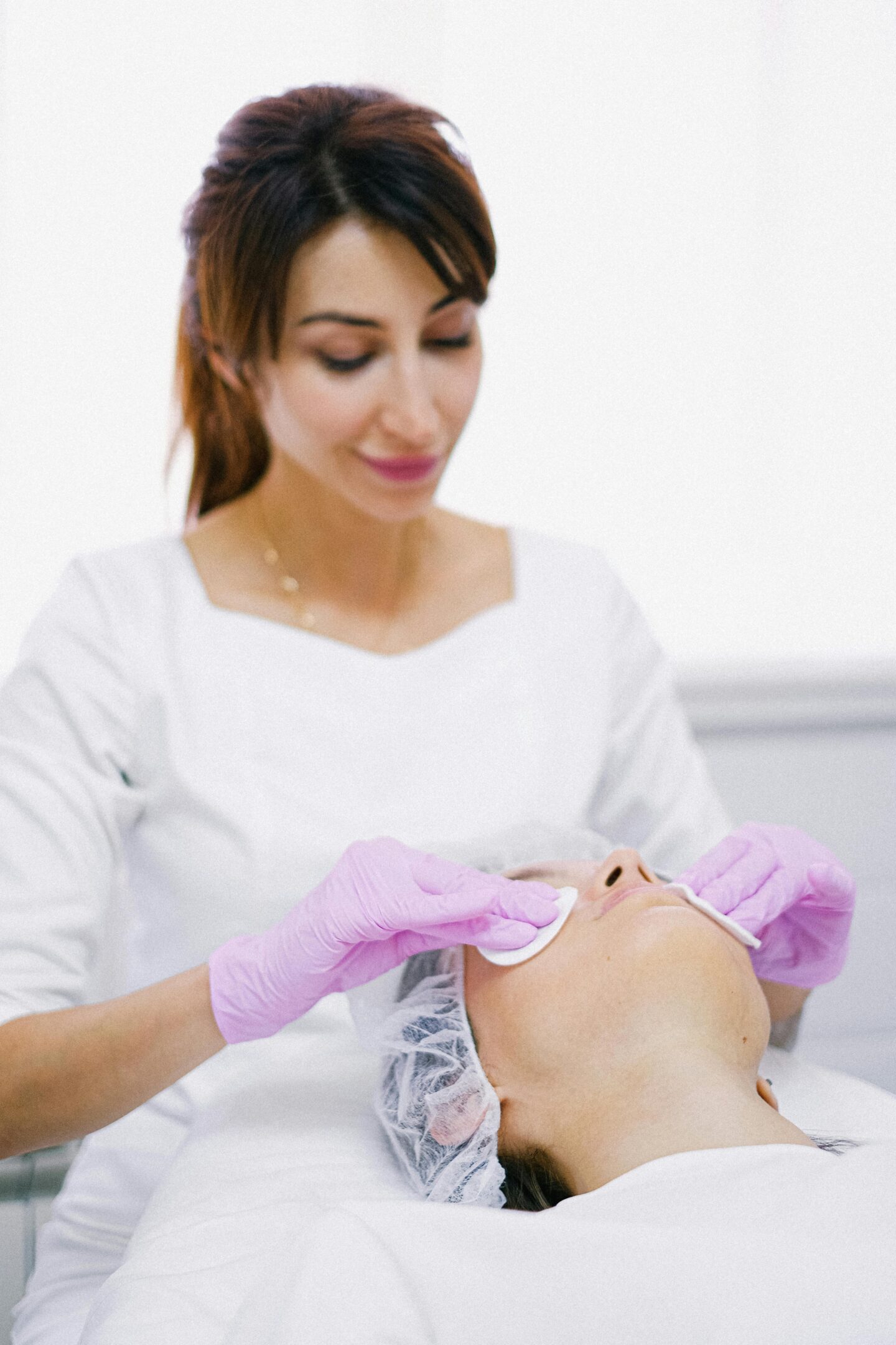 An esthetician wearing pink gloves cleans a client’s face with cotton pads in a skincare clinic.