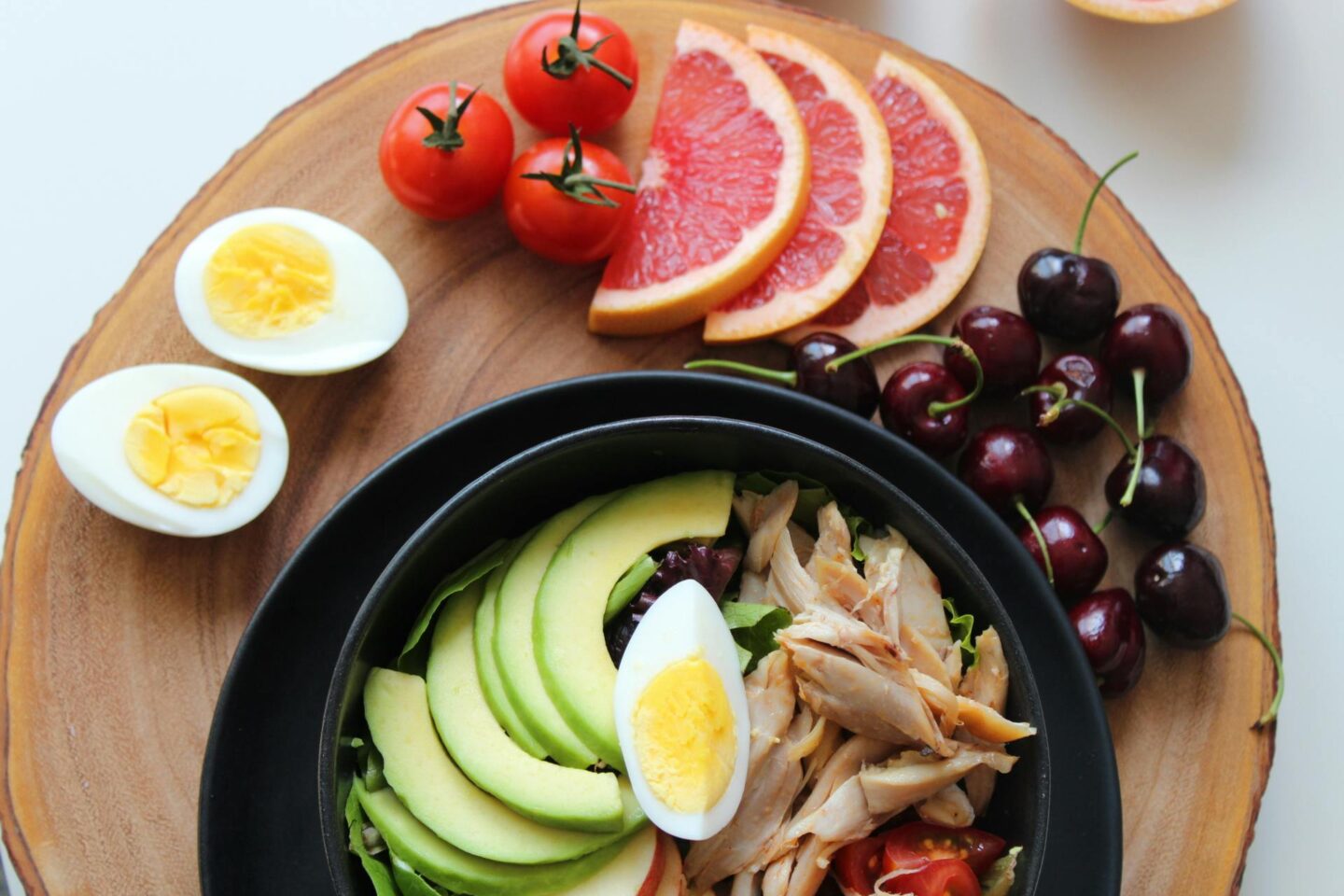 A healthy meal with avocado, eggs, chicken, tomatoes, grapefruit slices, and cherries arranged on a wooden plate.