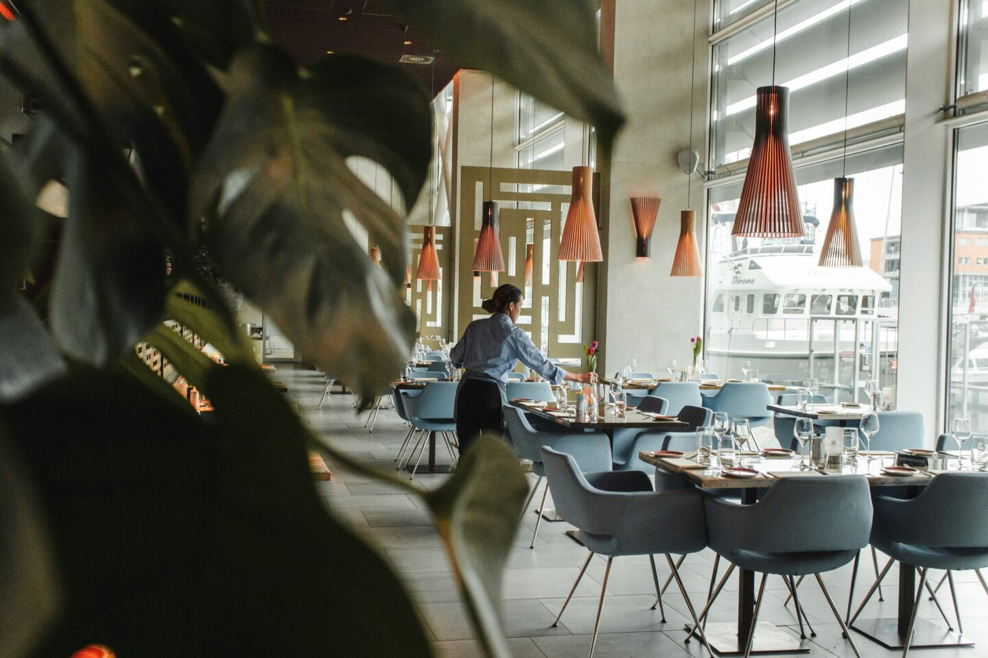 Restaurant interior with modern decor, blue chairs, and large windows overlooking a docked boat, as a staff member sets a table under warm pendant lights
