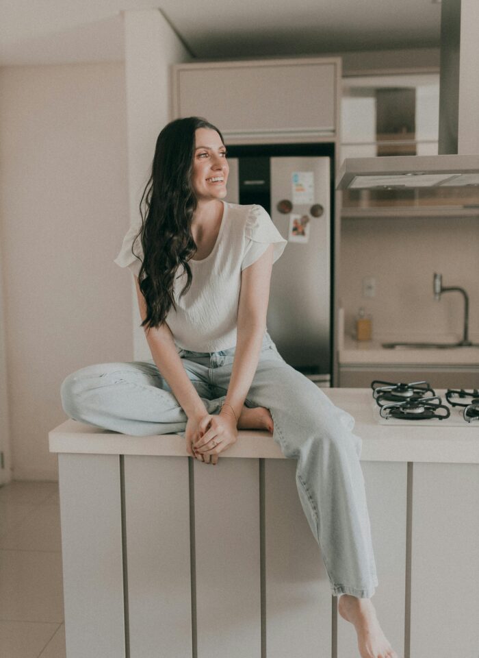 Woman sitting barefoot on a modern kitchen counter, smiling and looking to the side in a bright minimalist kitchen.