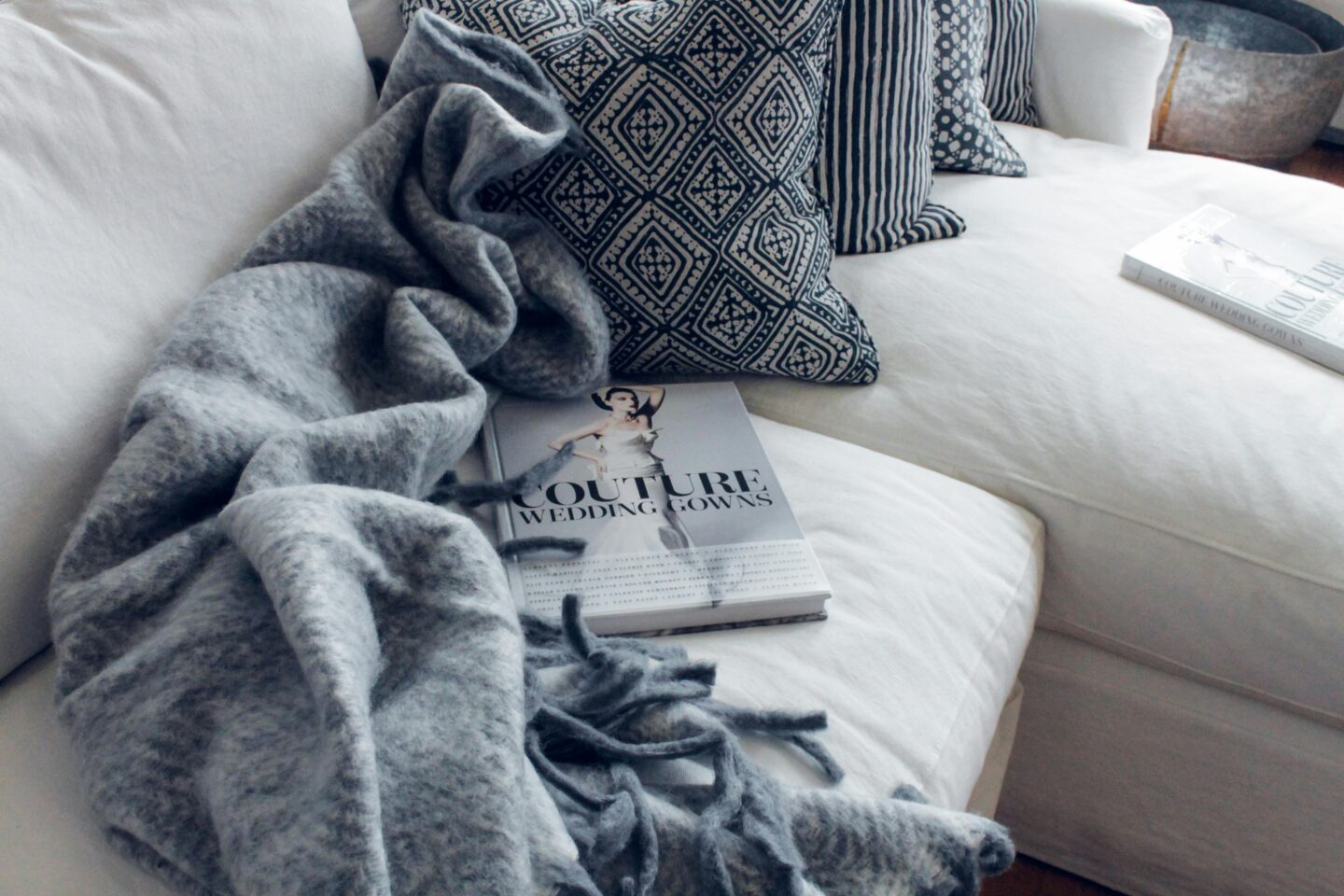 Cozy white sofa with blue patterned cushions, a soft gray blanket, and a coffee table book titled Couture Wedding Gowns.