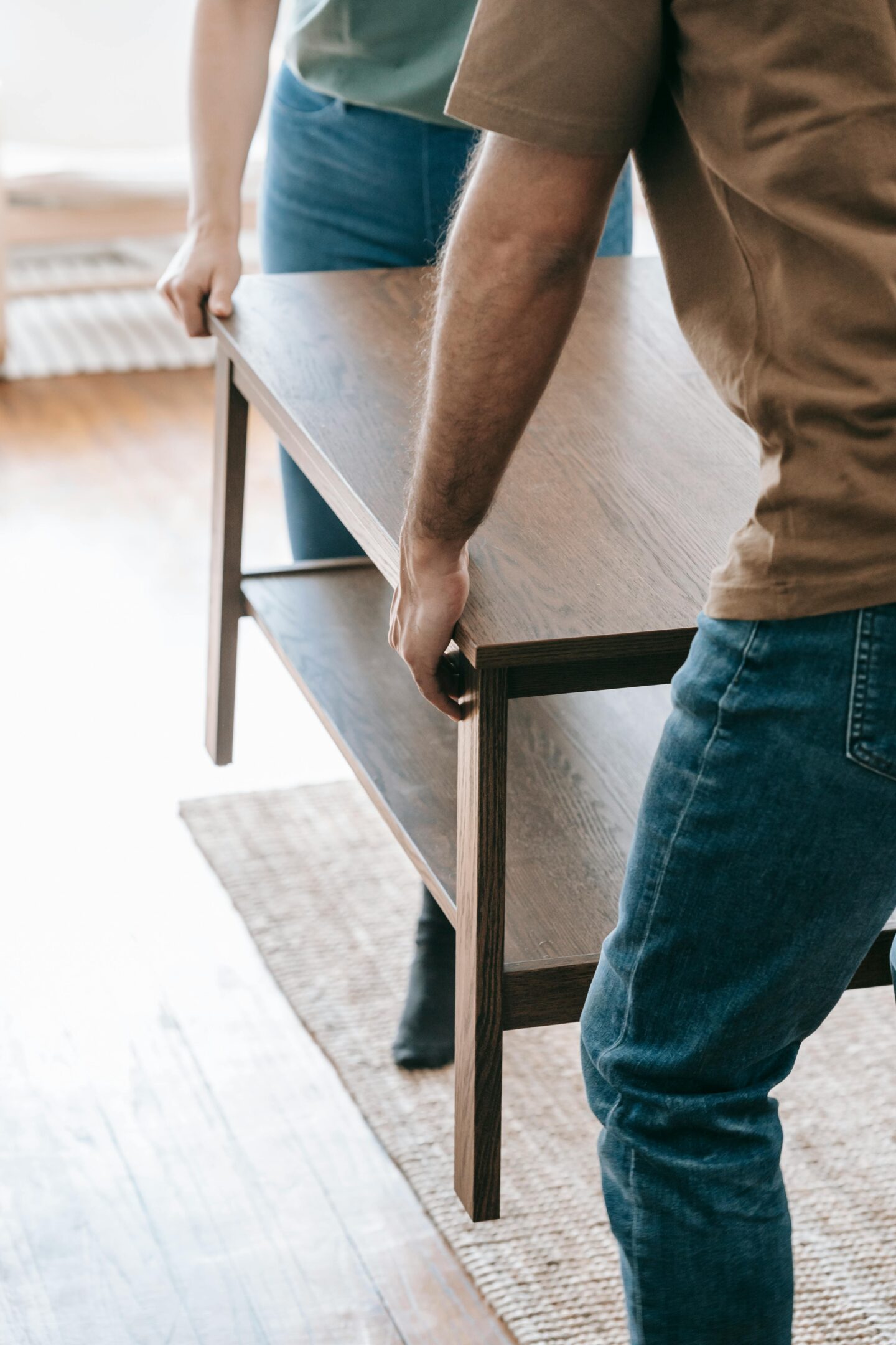 Two people lifting a wooden coffee table together in a bright living room with hardwood floors.