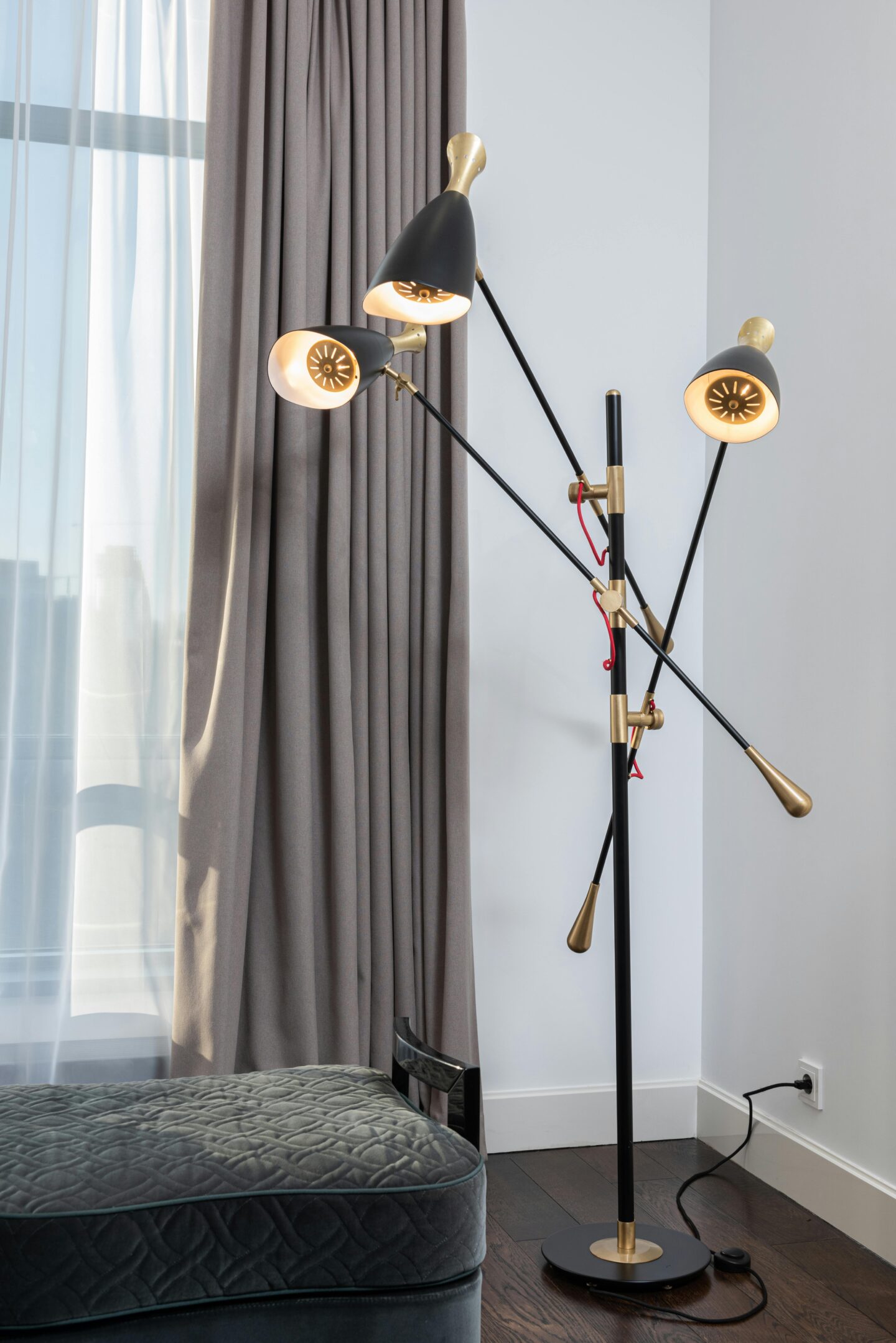 Modern black and gold floor lamp with three adjustable heads next to a gray curtain and velvet ottoman.