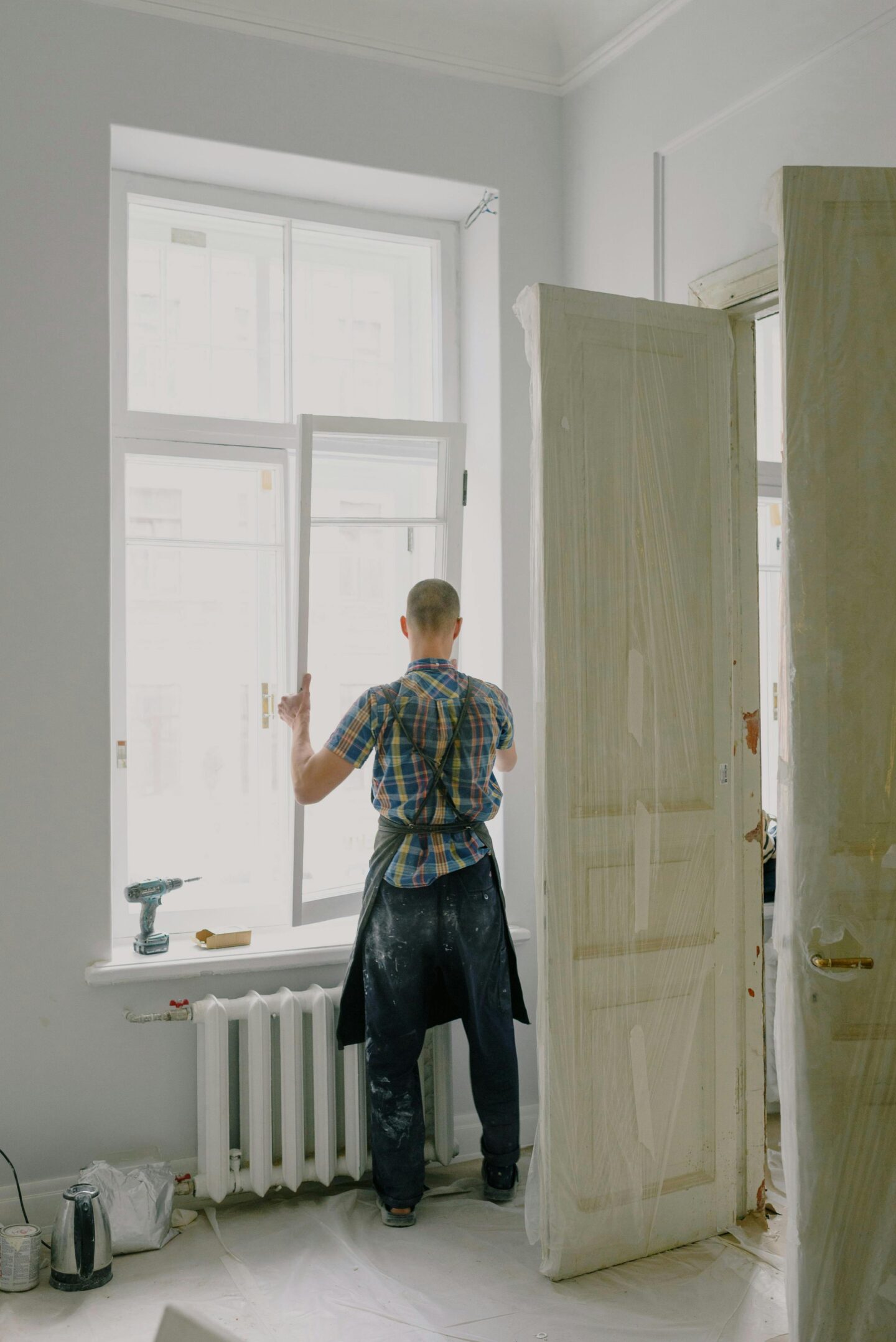 Man installing a window frame in a room undergoing renovation, with tools and protective coverings nearby.