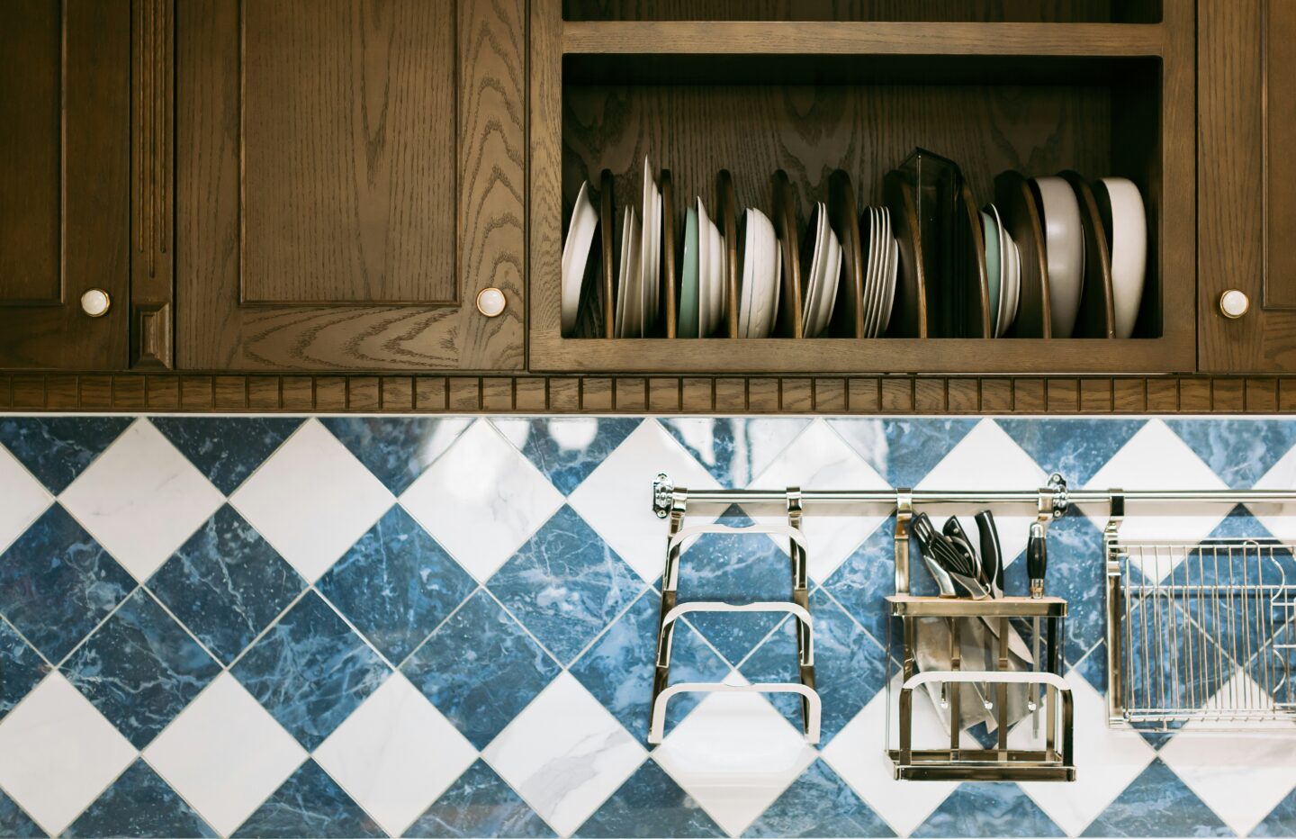 Wooden kitchen cabinets with neatly stacked plates above a blue and white checkered tile backsplash and stainless steel kitchen utensils hanging below.