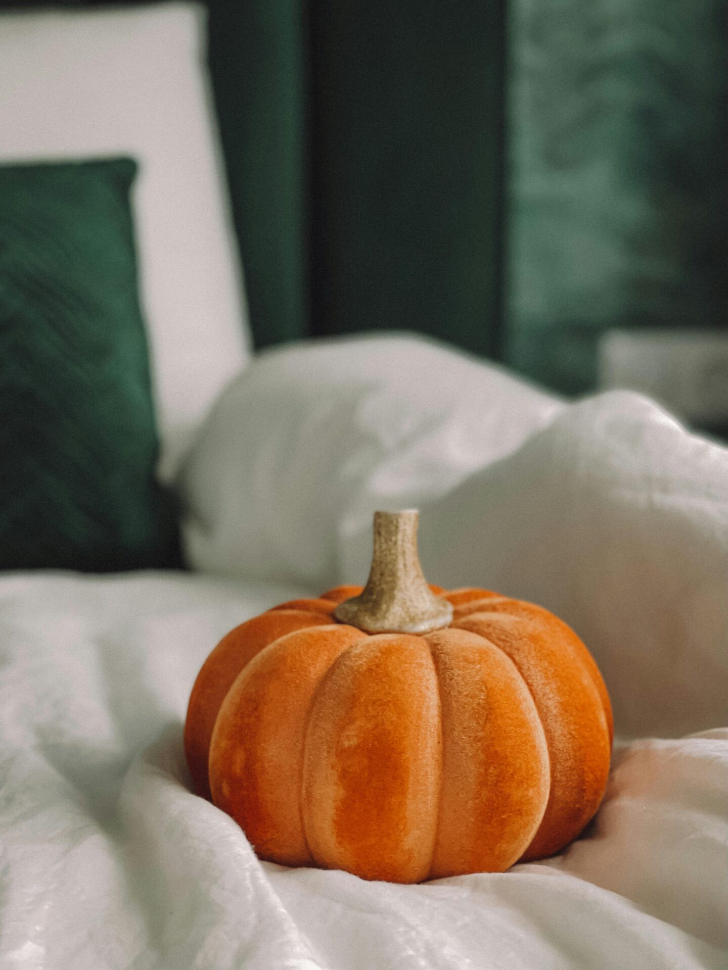 Soft orange velvet pumpkin placed on a white bed with green cushions in the background, creating a cozy autumn bedroom vibe.