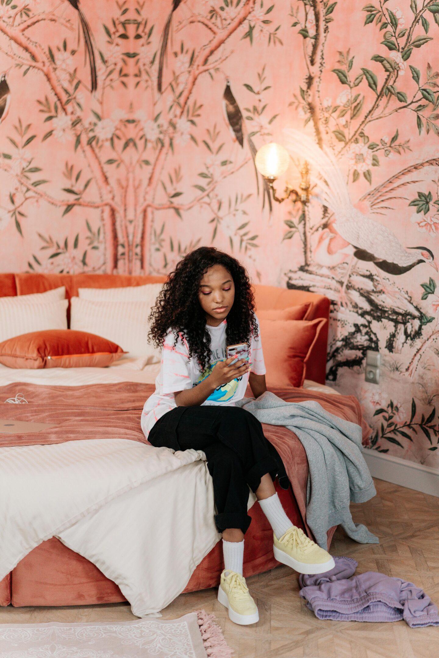 Young woman sitting on a bed with peach bedding and a pink botanical wallpaper featuring cranes, using her phone in a stylish bedroom.