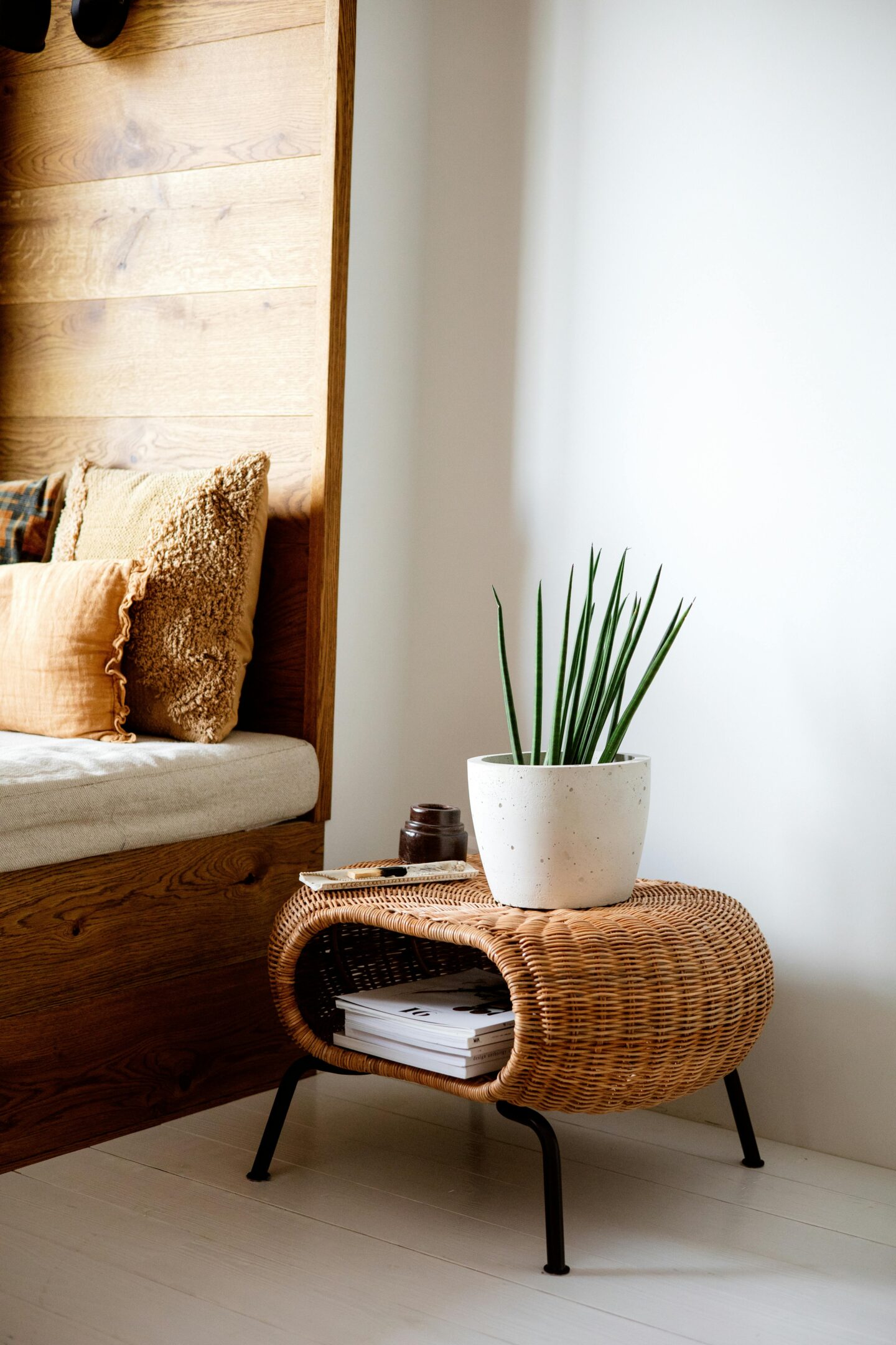Cozy corner with neutral-toned cushions, a woven rattan side table holding a white planter with a green plant, and neatly stacked magazines underneath.