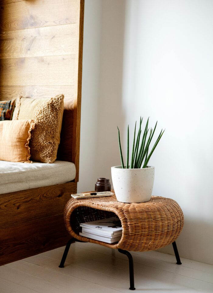 Cozy corner with neutral-toned cushions, a woven rattan side table holding a white planter with a green plant, and neatly stacked magazines underneath.