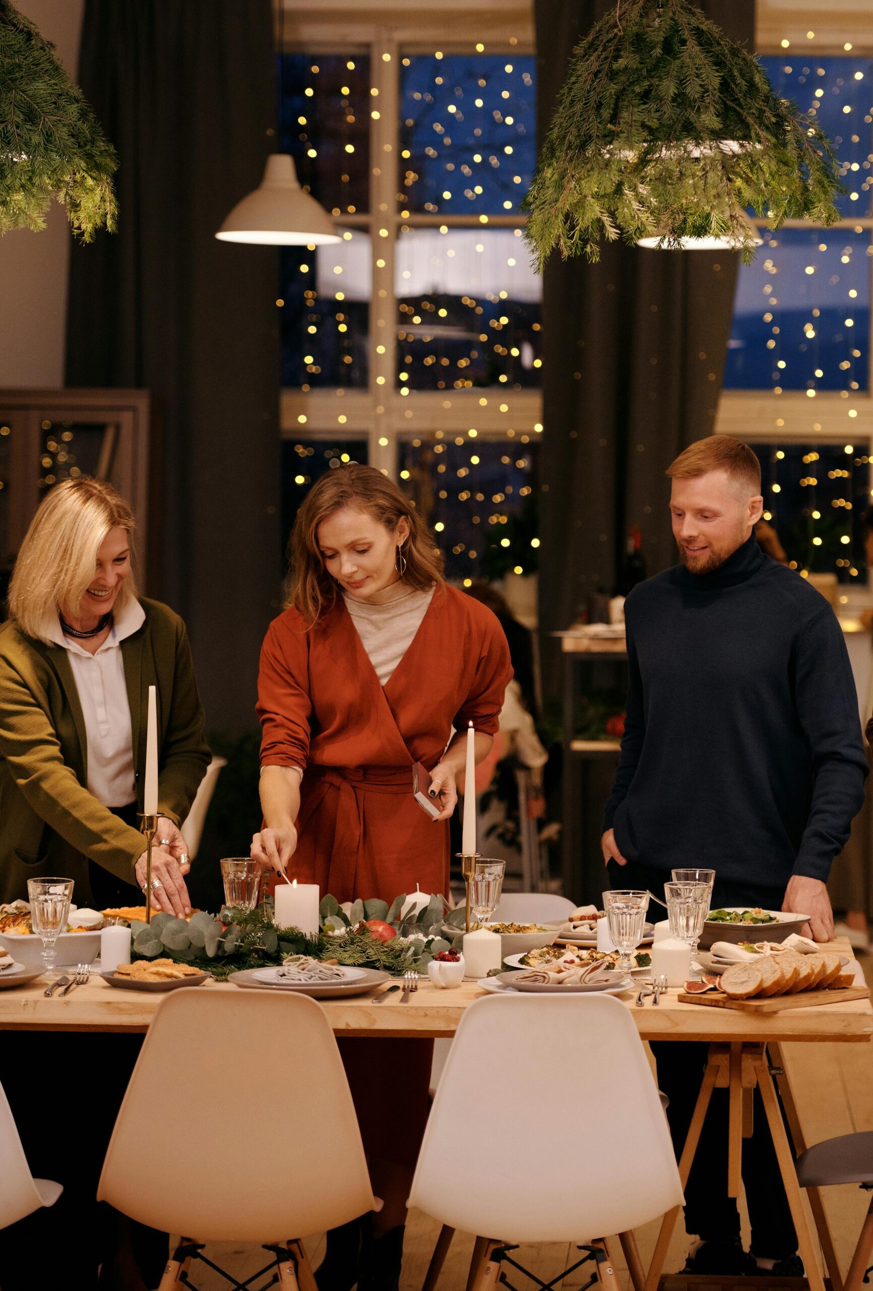 Three people setting a festive dinner table decorated with candles, greenery, and holiday lights glowing in the background by a large window.