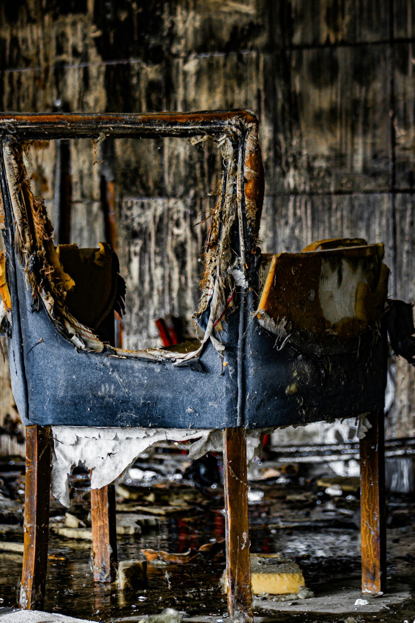 Burnt and decaying chair with torn upholstery in an abandoned, soot-covered room.