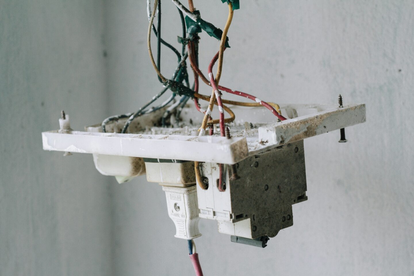 Close-up of exposed and tangled electrical wires attached to a dirty, damaged switchboard on a wall.