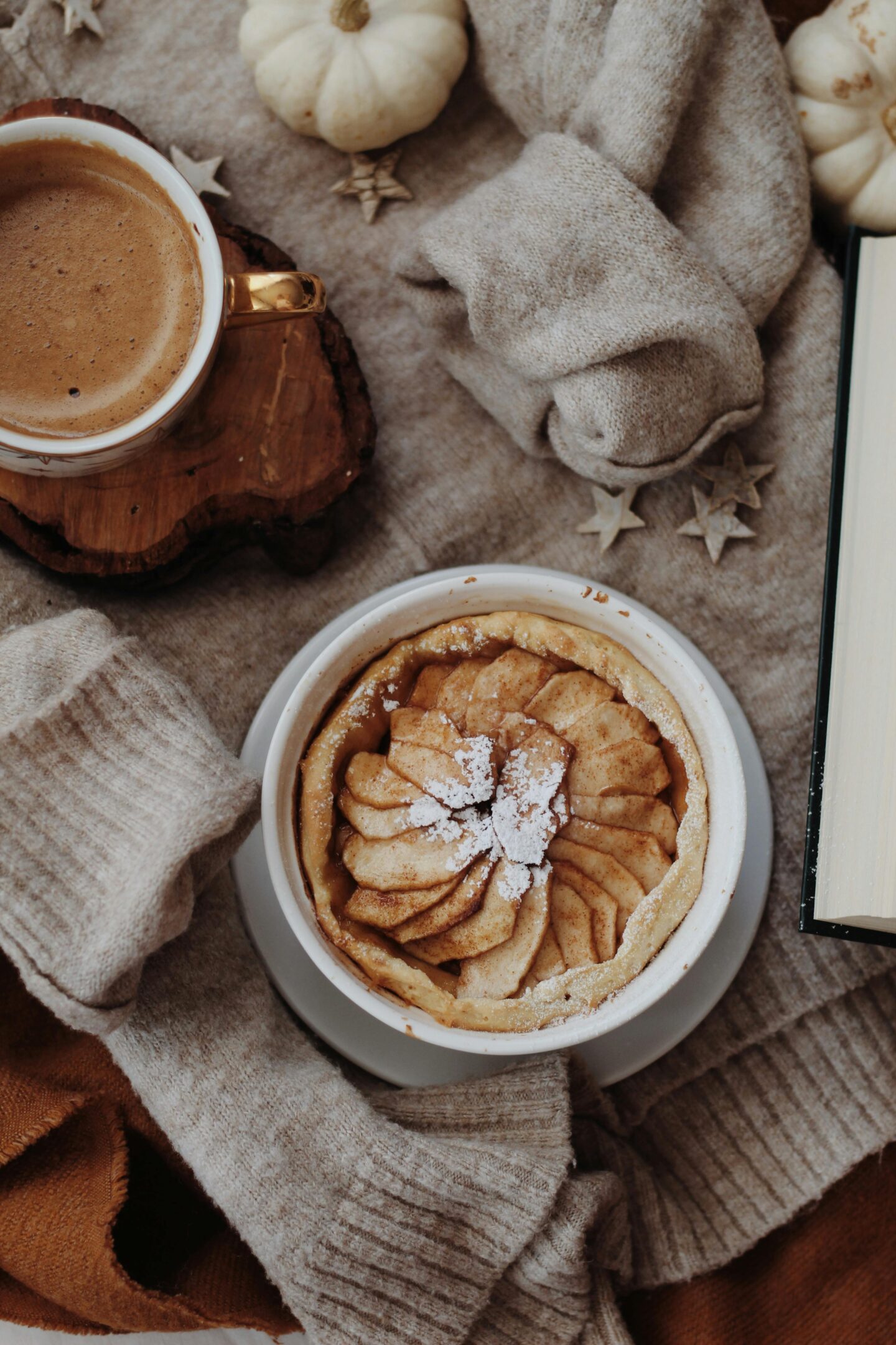 Cozy winter scene with a warm drink and an apple tart topped with powdered sugar, surrounded by soft knits and seasonal decor.