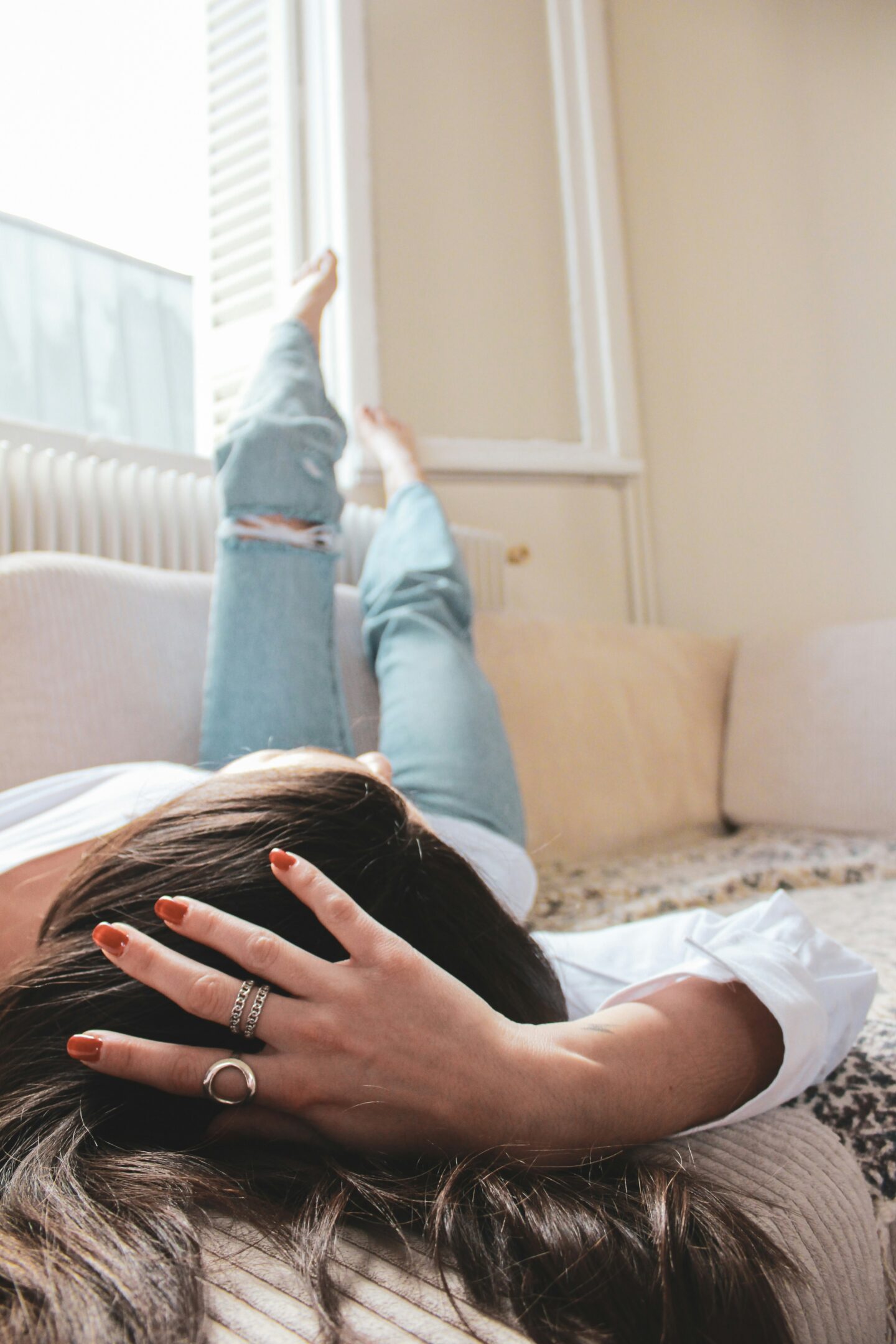 Woman relaxing on a sofa with her legs up by the window, wearing jeans and showcasing silver rings on her hand.