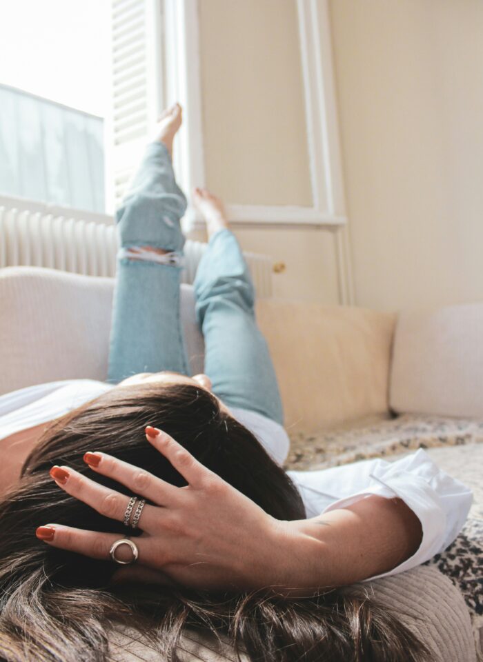 Woman relaxing on a sofa with her legs up by the window, wearing jeans and showcasing silver rings on her hand.