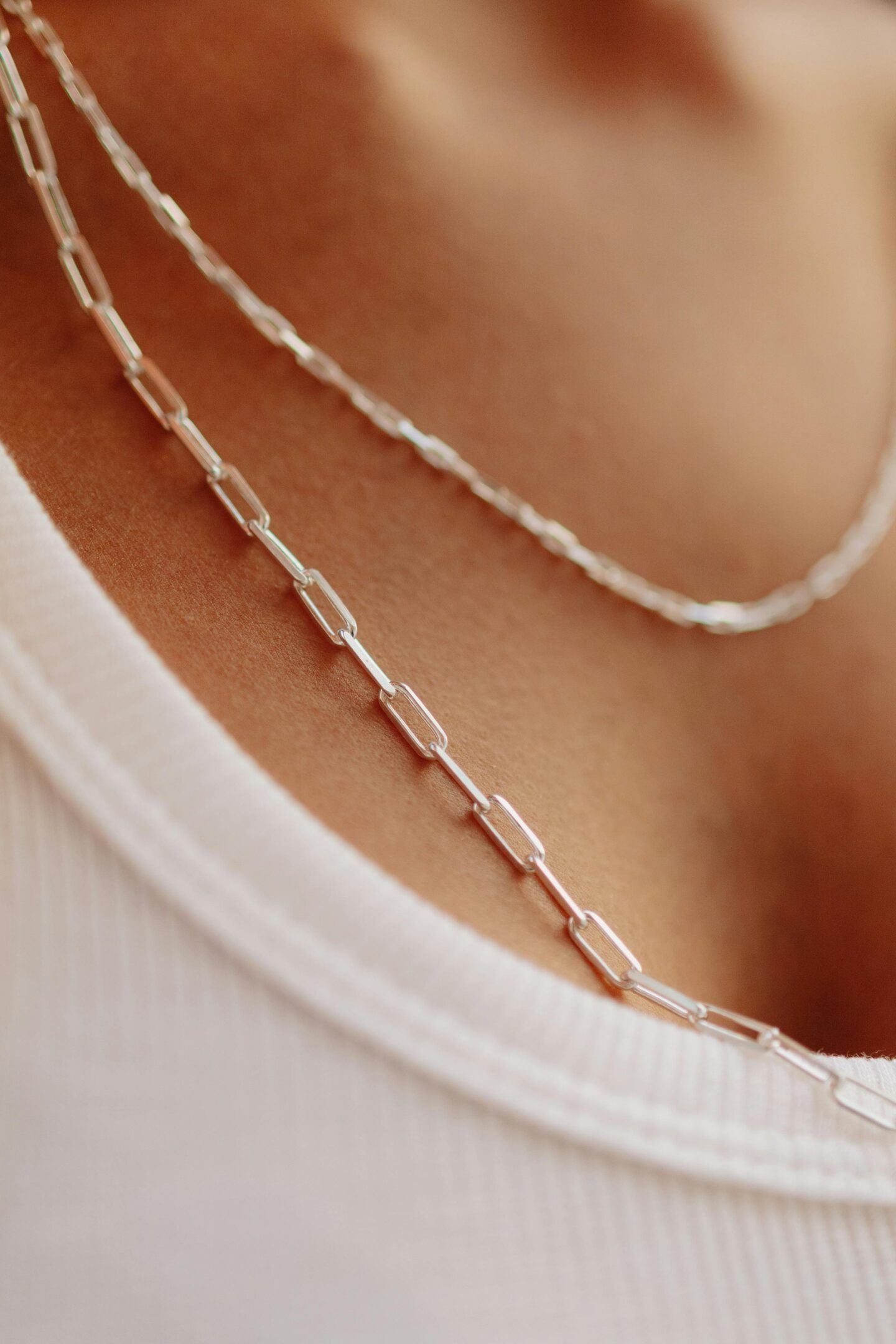 Close-up of layered paperclip chain silver necklaces with fine rectangular links resting on the collarbone above a white top.