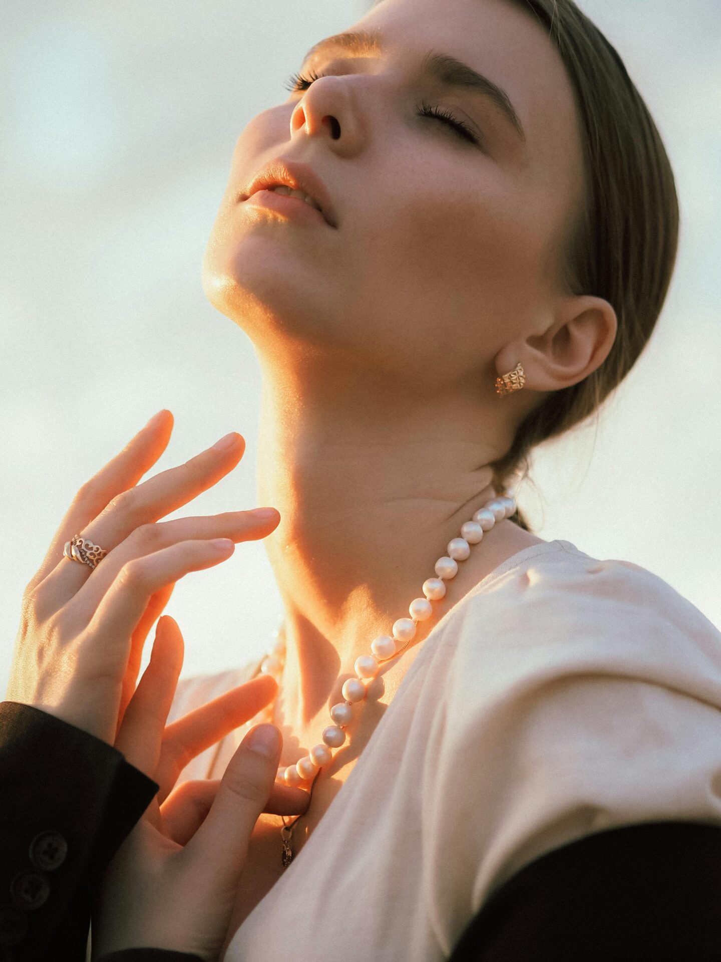 Woman bathed in golden light wearing a pearl necklace, gold earrings, and rings, exuding classic elegance.