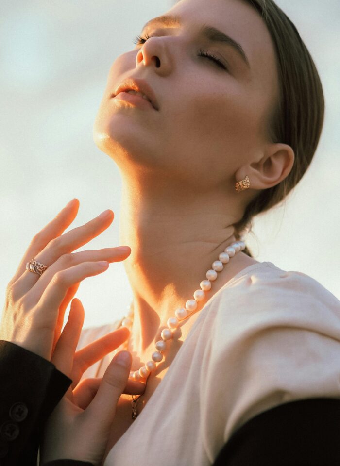 Woman bathed in golden light wearing a pearl necklace, gold earrings, and rings, exuding classic elegance.