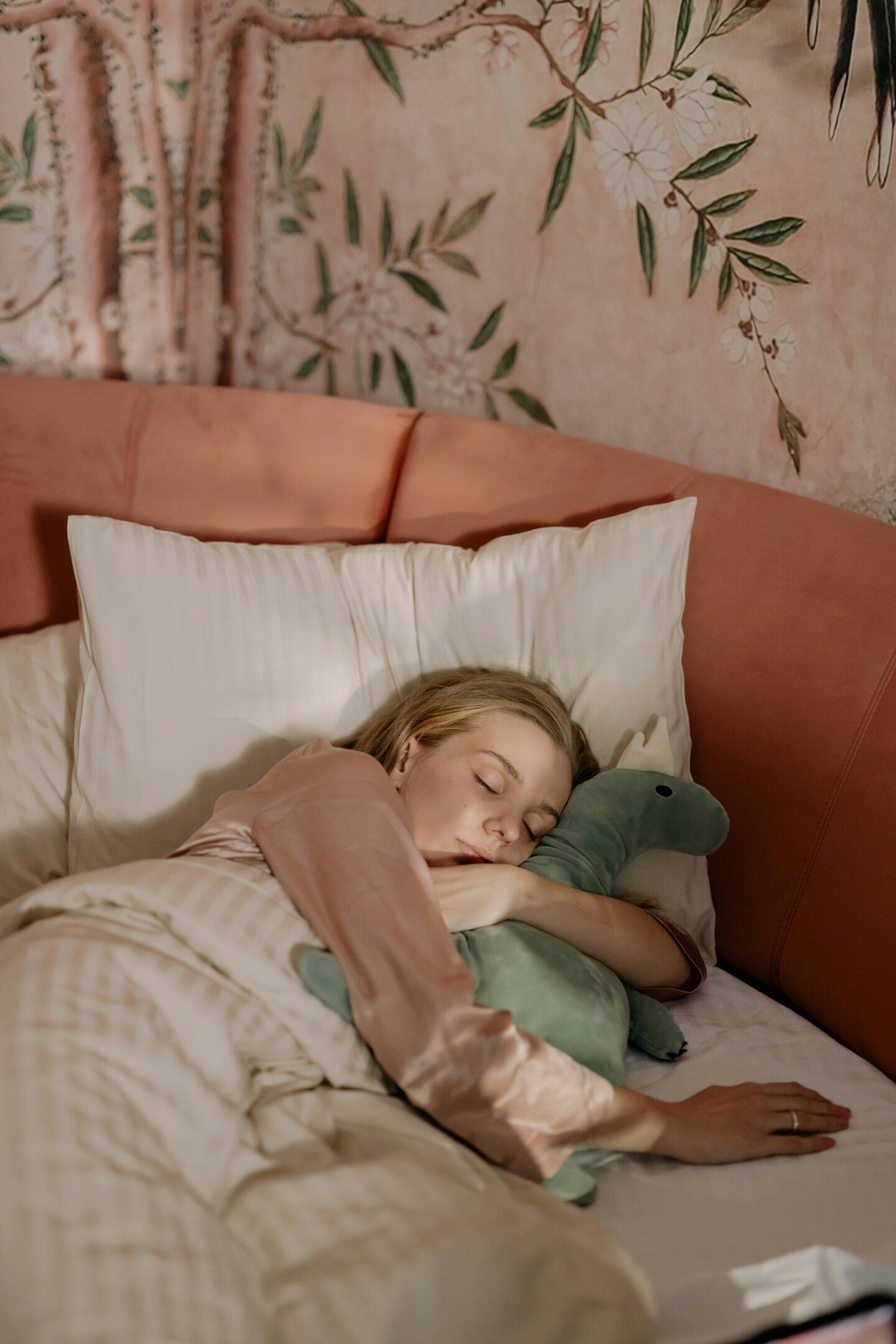 Young woman peacefully sleeping in bed, hugging a green plush dinosaur, with a soft pink headboard and floral wallpaper in the background.