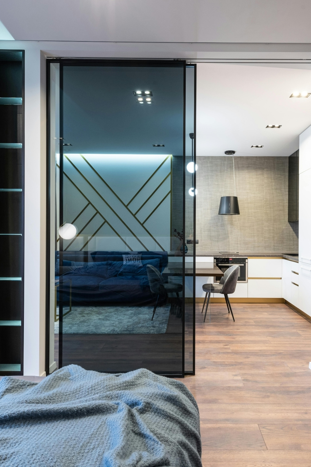 Modern open-plan apartment with a glass sliding door separating the bedroom from the kitchen and living area, featuring minimalist decor, wooden floors, and geometric wall design with LED lighting.
