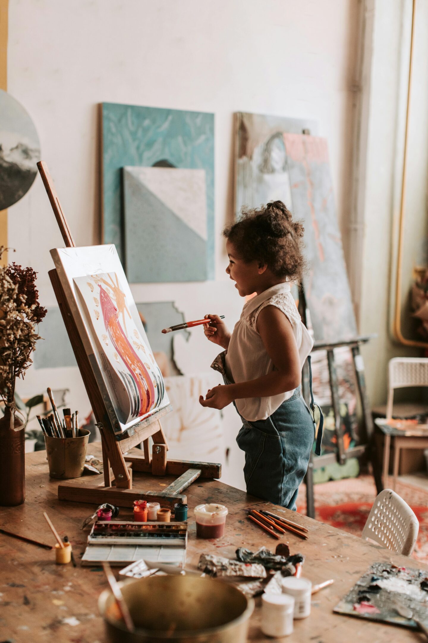 Young girl smiling while painting on an easel in an art studio surrounded by brushes and paint supplies.