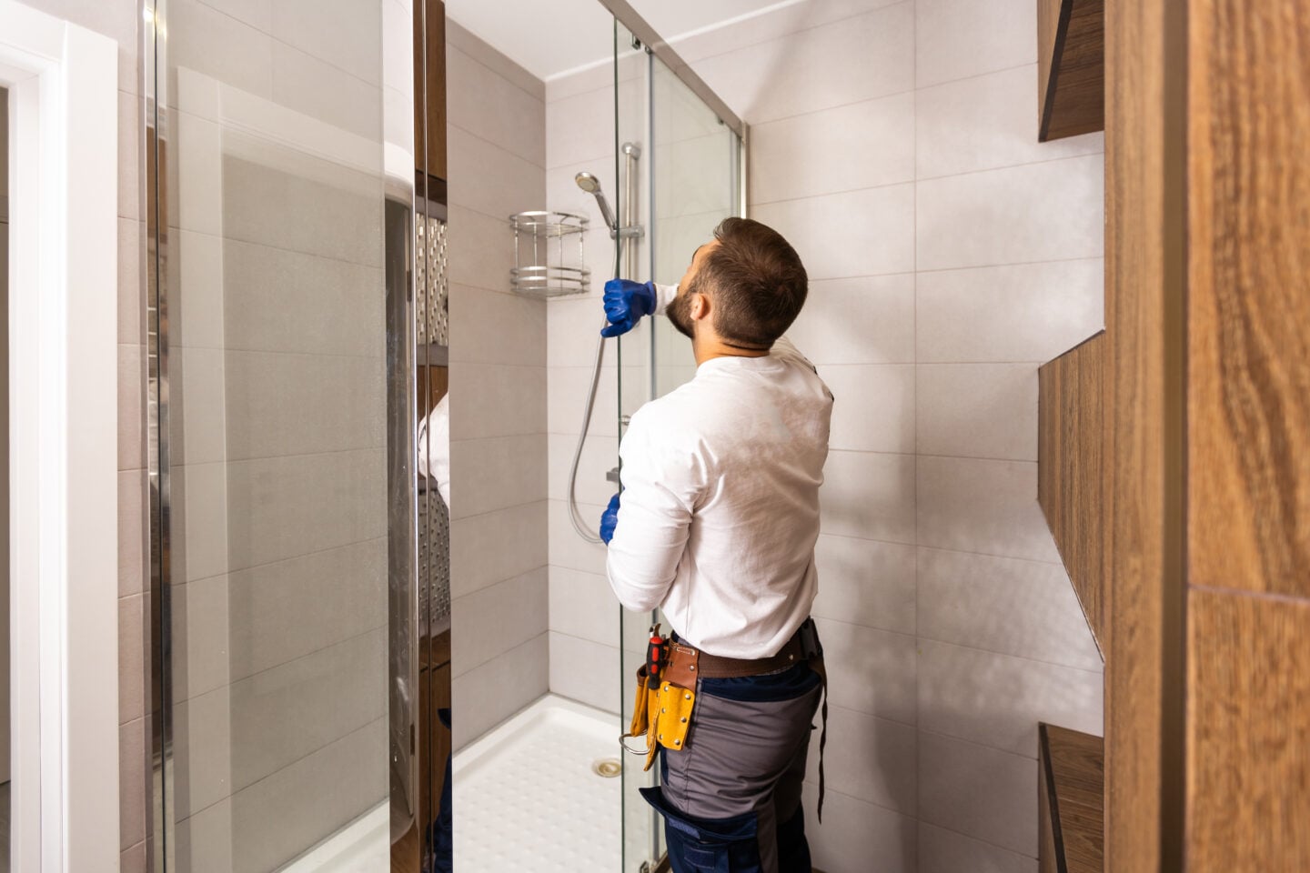 A manwearing gloves and a tool belt adjusts a handheld shower inside a modern bathroom.