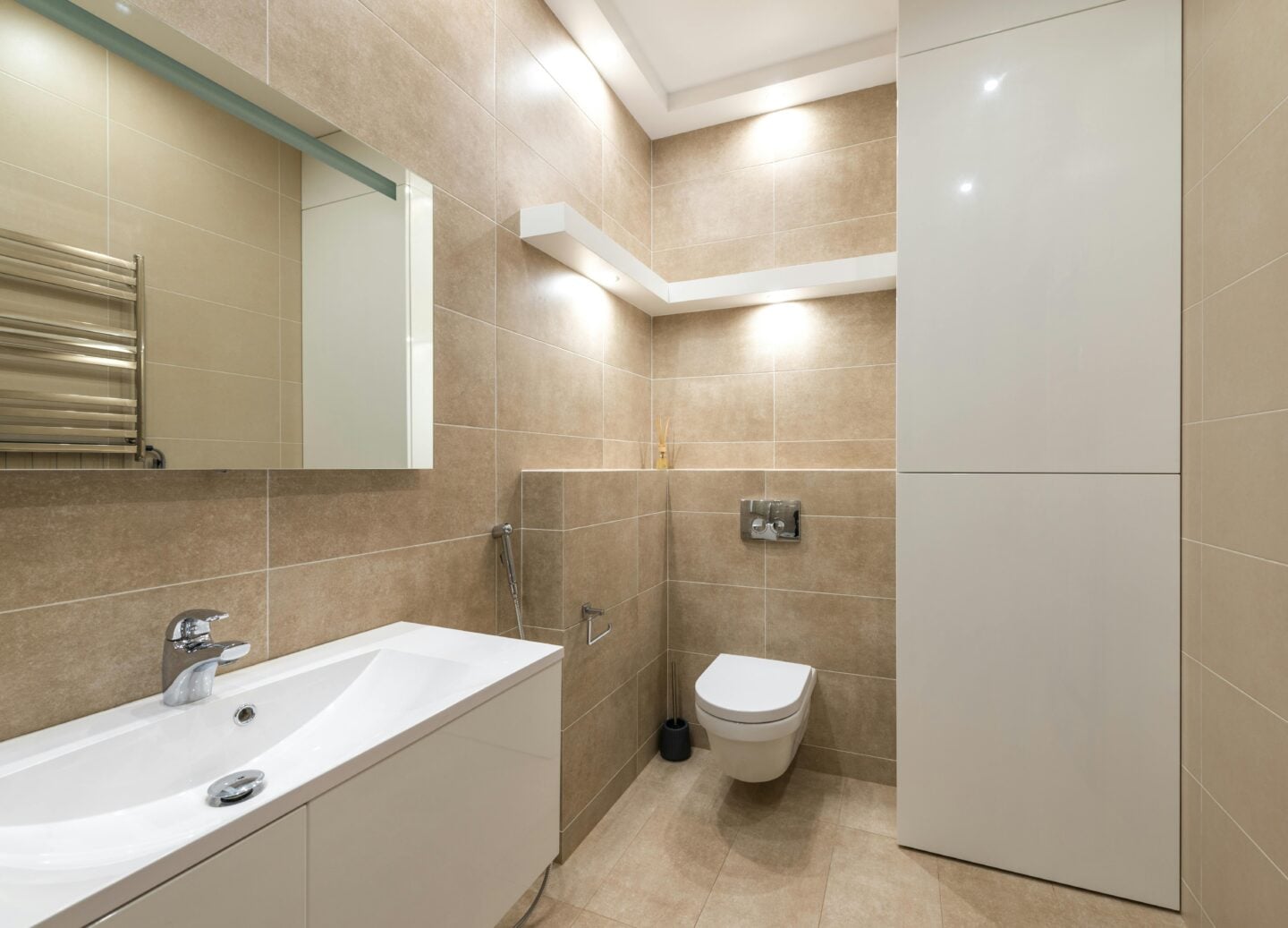 A beige-tiled bathroom with a floating sink, wall-mounted toilet, light fixtures and built-in storage cabinet.