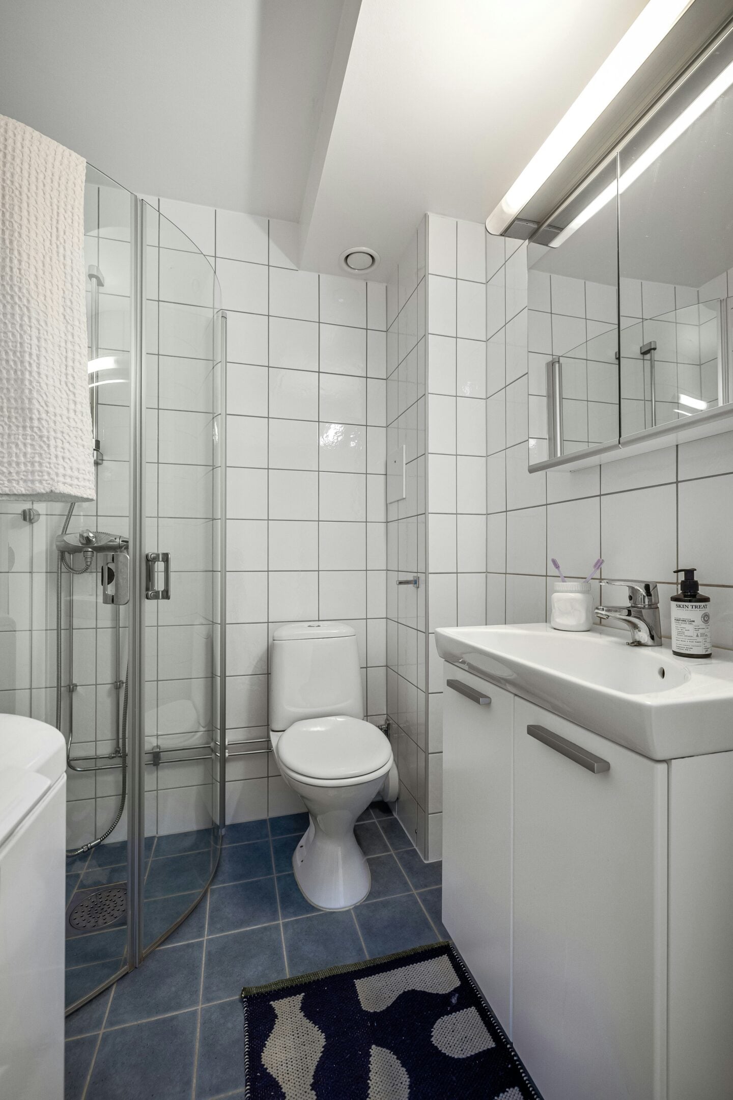 A compact bathroom with white tiled walls, a glass shower enclosure, a toilet, and a white vanity with a mirror cabinet.