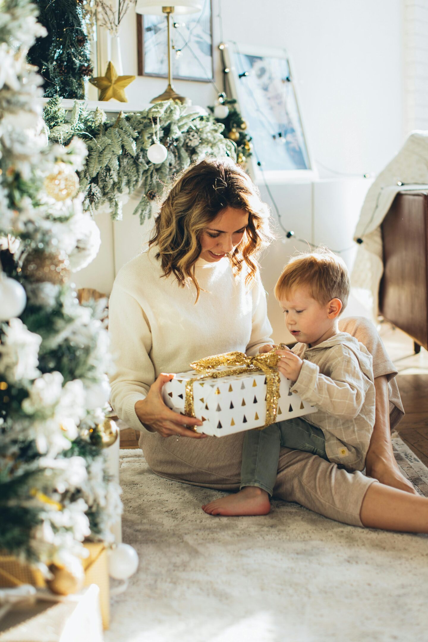 Mother and young son sitting by a Christmas tree, unwrapping a white gift box with gold ribbon.