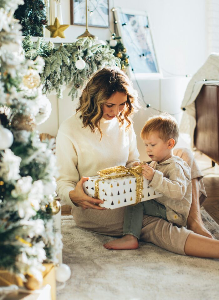 Mother and young son sitting by a Christmas tree, unwrapping a white gift box with gold ribbon.