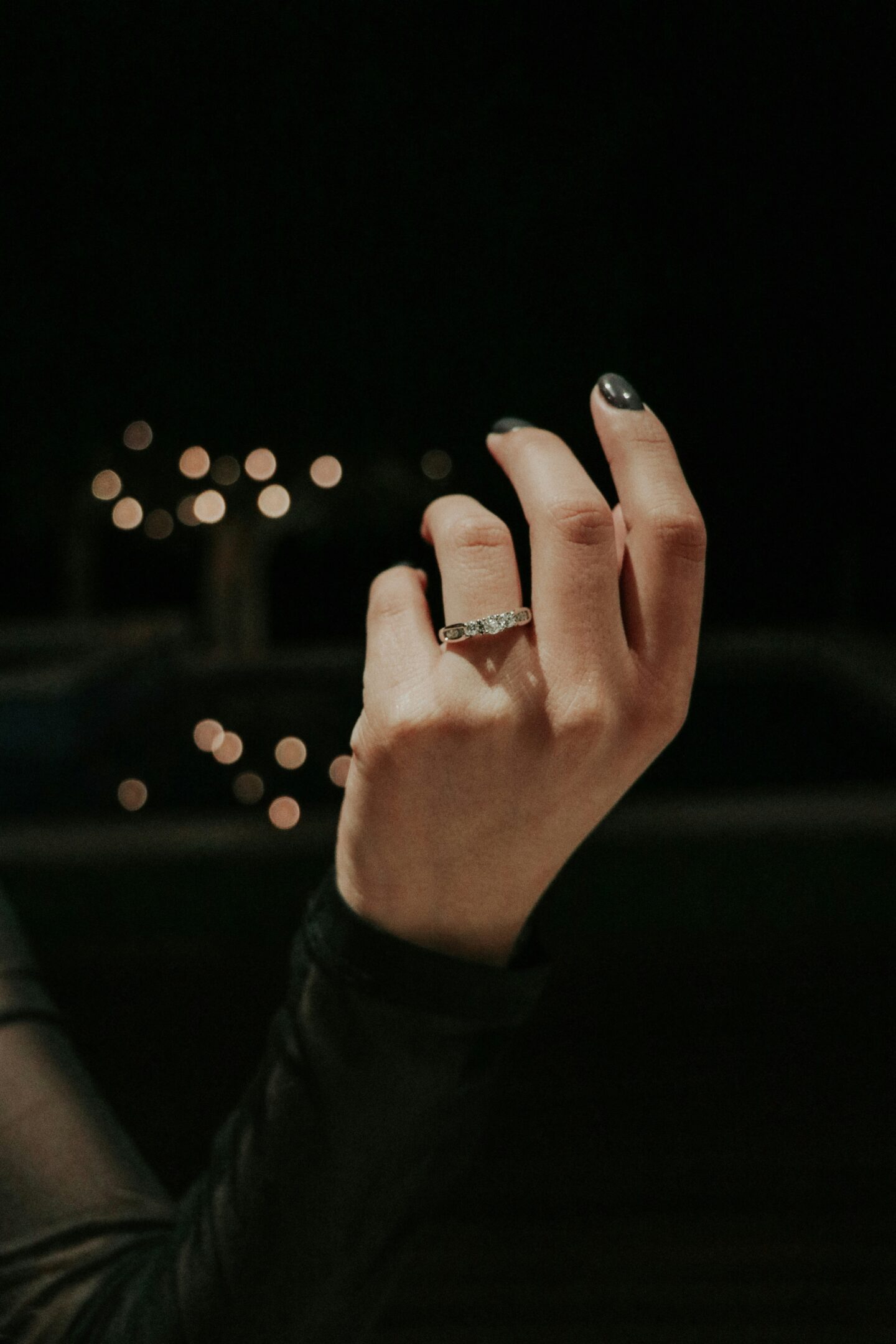 Close-up of a hand with dark nail polish wearing a diamond engagement ring against a dark background with blurred golden lights.