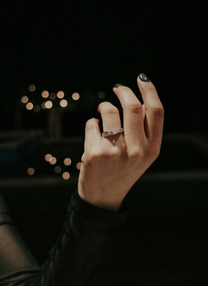Close-up of a hand with dark nail polish wearing a diamond engagement ring against a dark background with blurred golden lights.