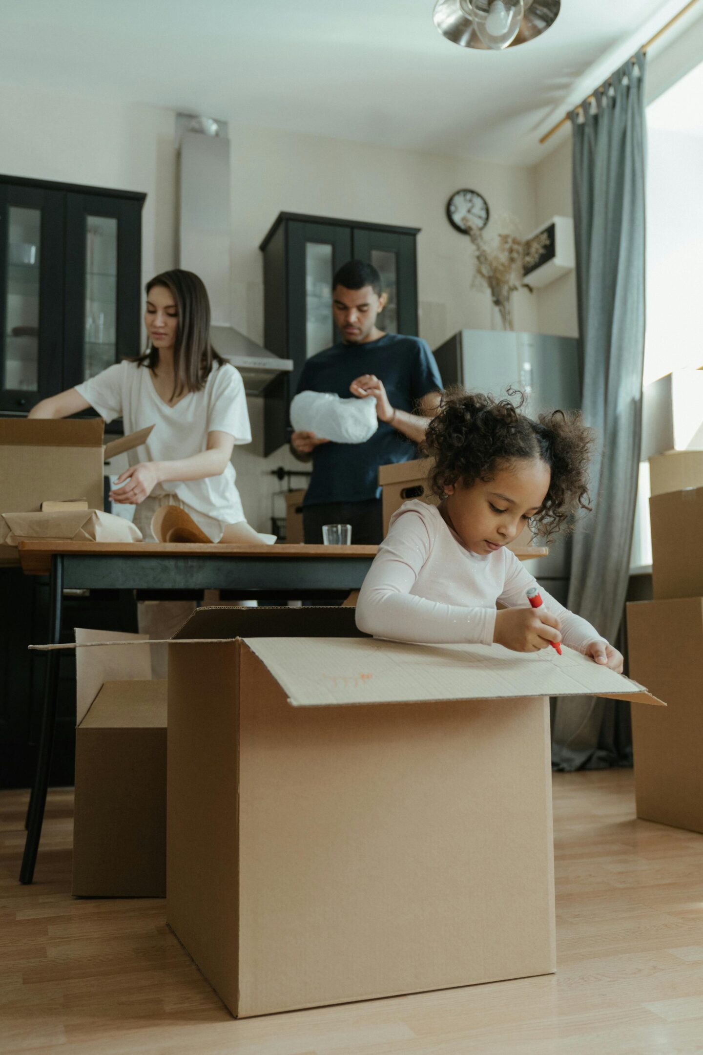 A young girl drawing on a cardboard box while two adults pack items in the background of a kitchen.
