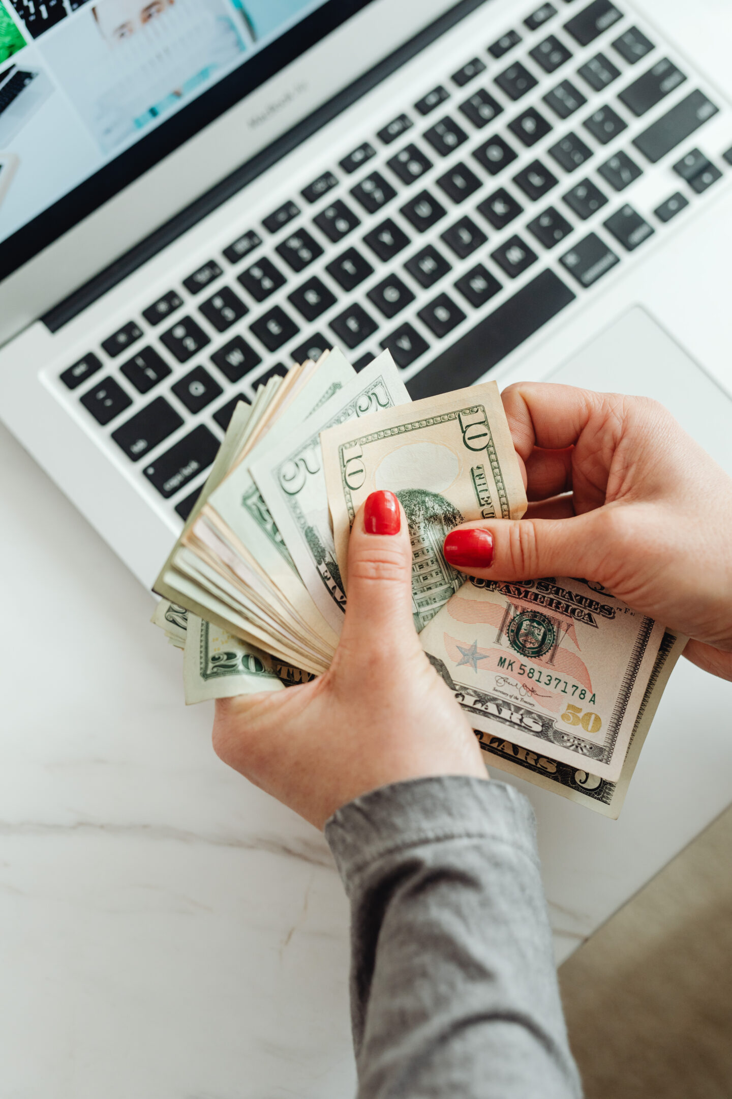 Person with red nail polish counting U.S. dollar bills in front of an open laptop on a white desk.